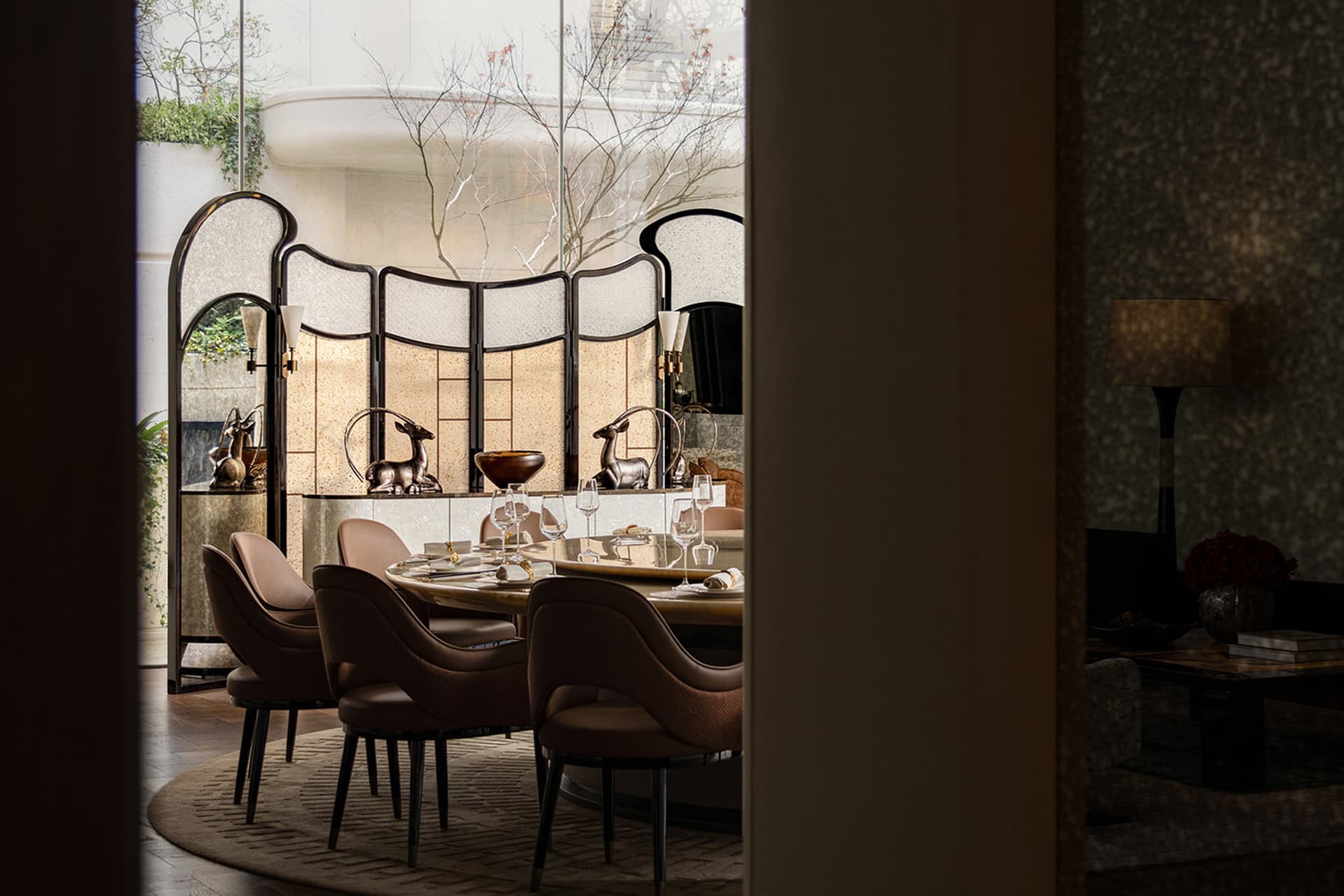 A modern dining room with a large wooden dining table and chairs, set with various items, surrounded by a large window allowing natural light to fill the space