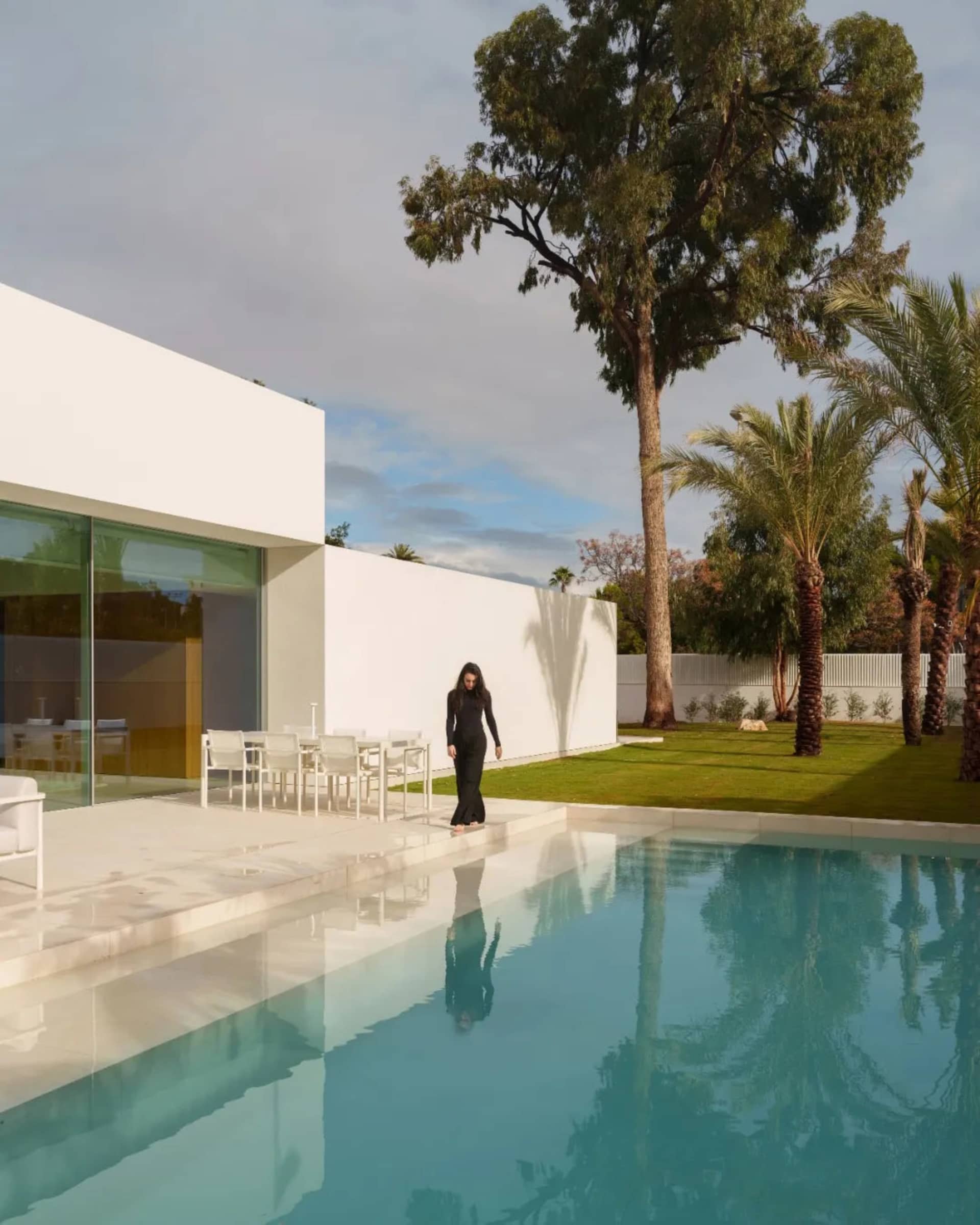 A modern outdoor living space with a pool surrounded by a white wall, a patio with a dining table and chairs, and potted plants, with a person standing near the pool, lit by natural sunlight