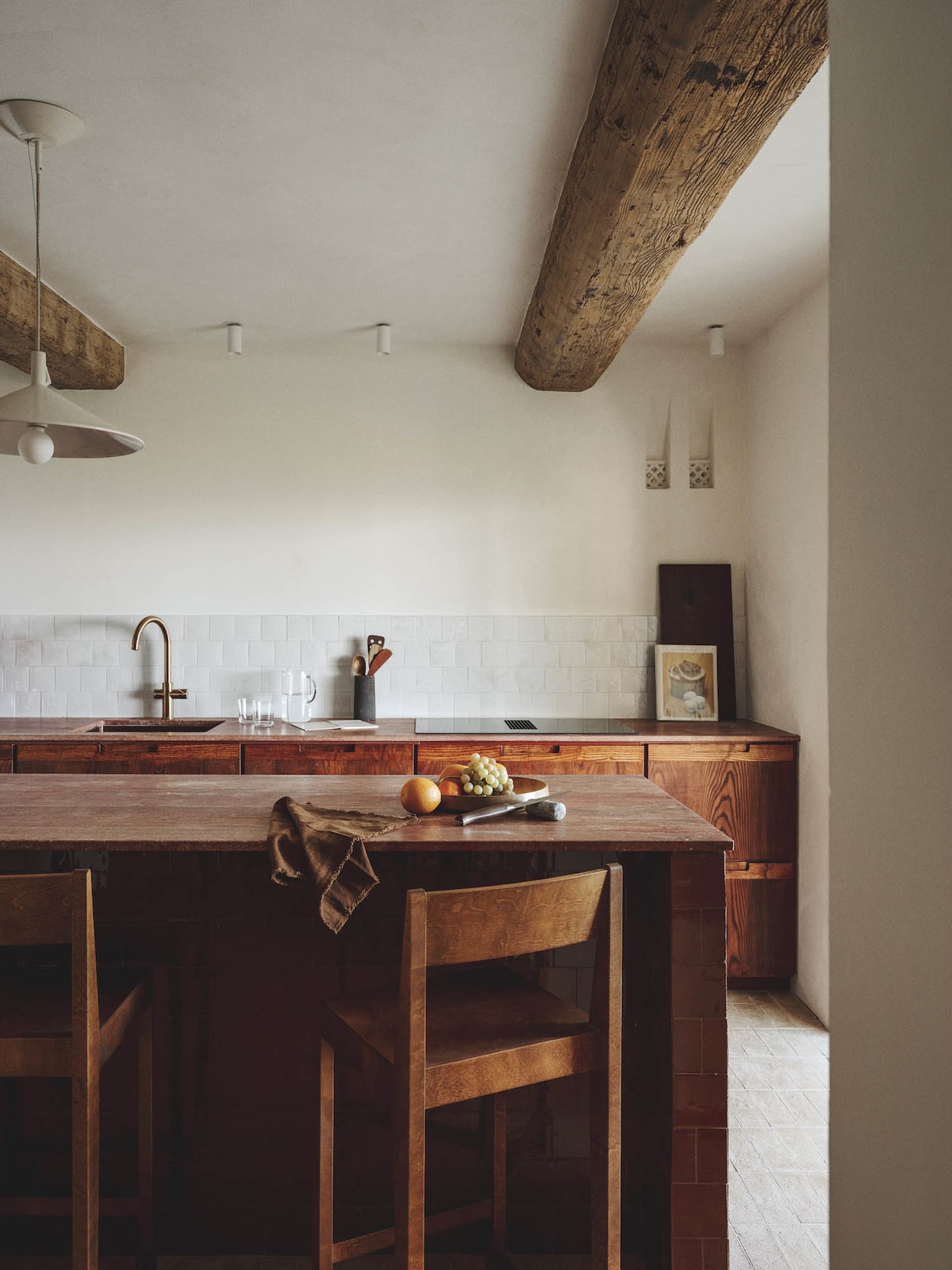 Modern kitchen with a wooden countertop, dining table, and chairs, featuring a sink, refrigerator, and bowl, with a wooden floor and tiled wall, lit by natural sunlight