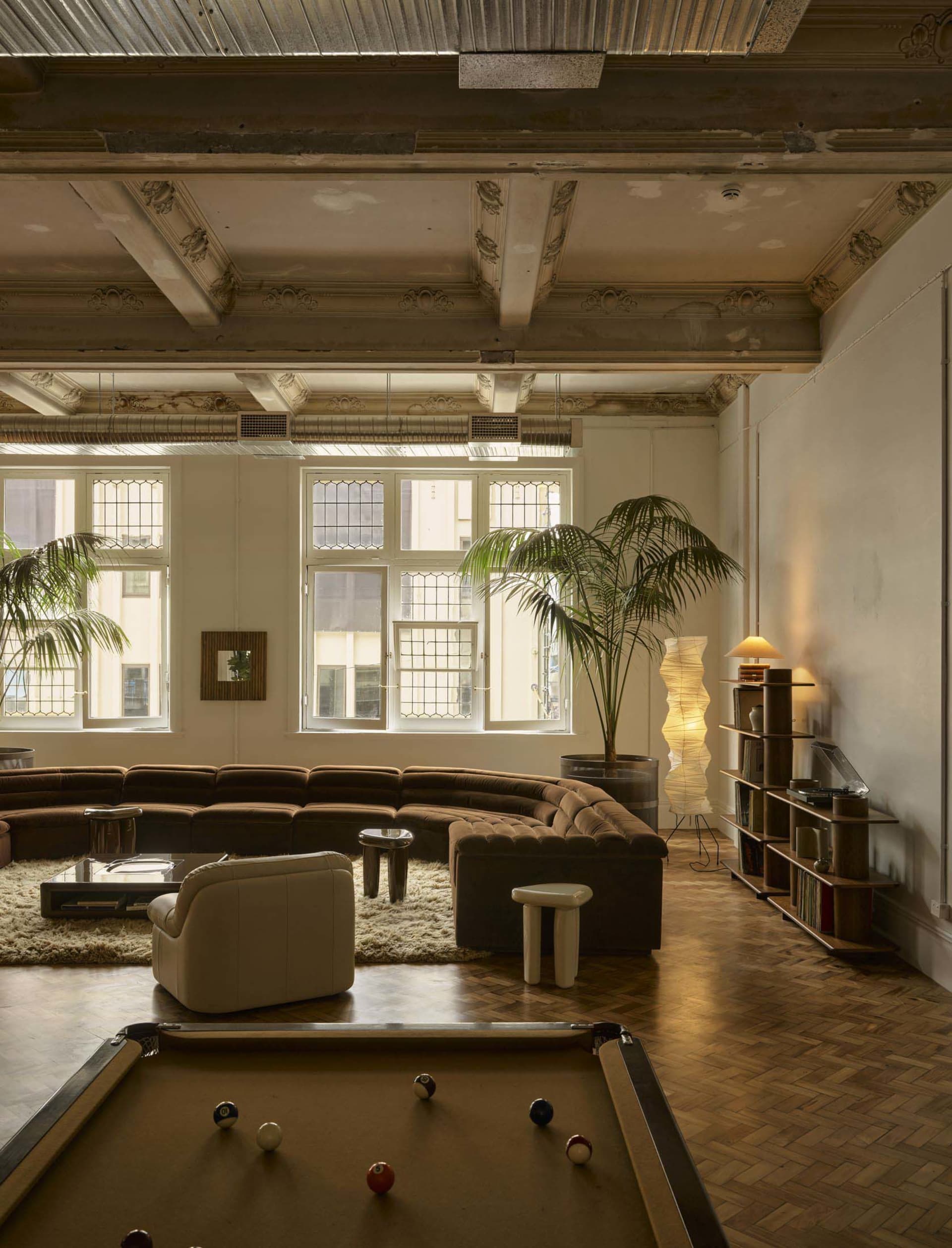 A modern living room with a pool table, large couch, chair, dining table, and potted plants, filled with natural light from multiple windows