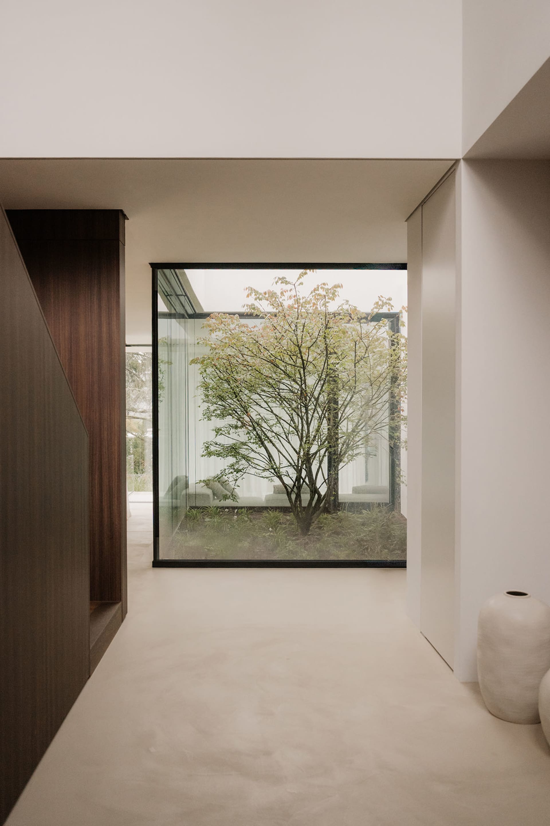 A living room with a couch, chair, and wooden floor, featuring a large glass window with a tree outside, allowing natural light to enter and creating a bright and welcoming atmosphere