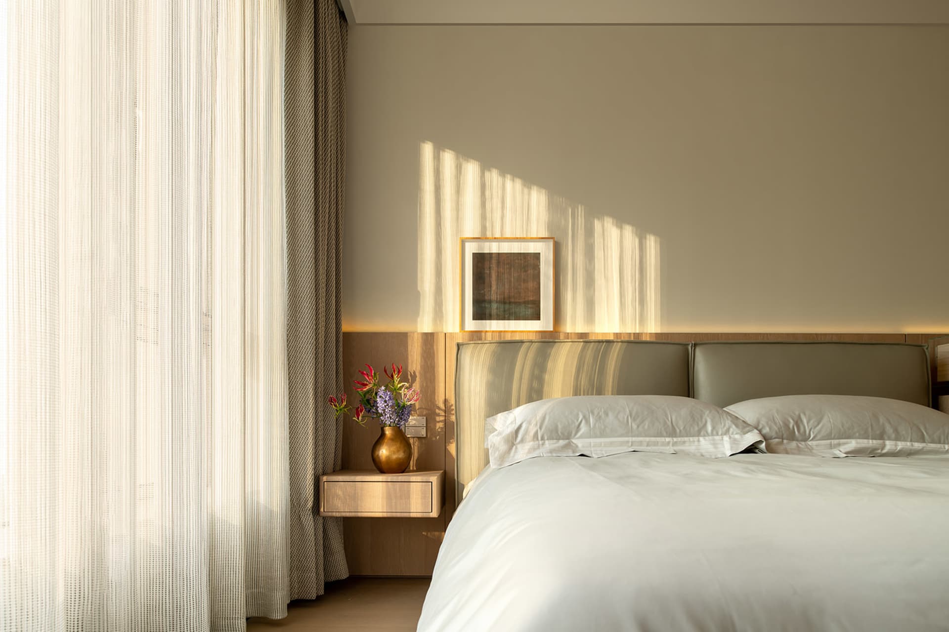 A modern bedroom with a bed, a vase with flowers, and a picture frame on the nightstand, set against a predominantly white background with natural lighting