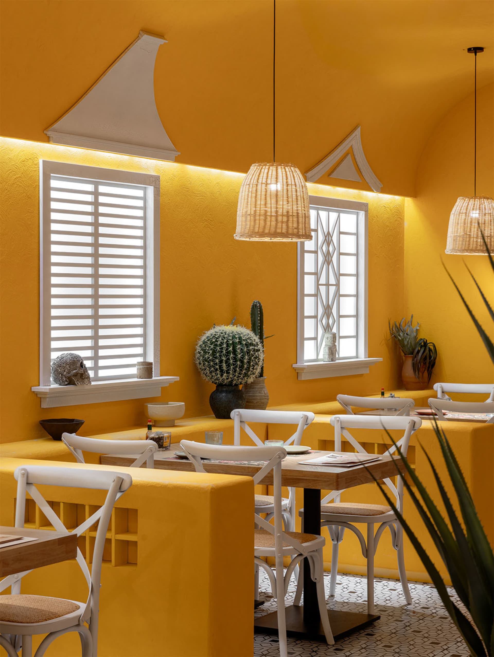 Cozy modern restaurant with a yellow wall, white furniture, and potted plants, warm lighting, and a dining area with tables and chairs