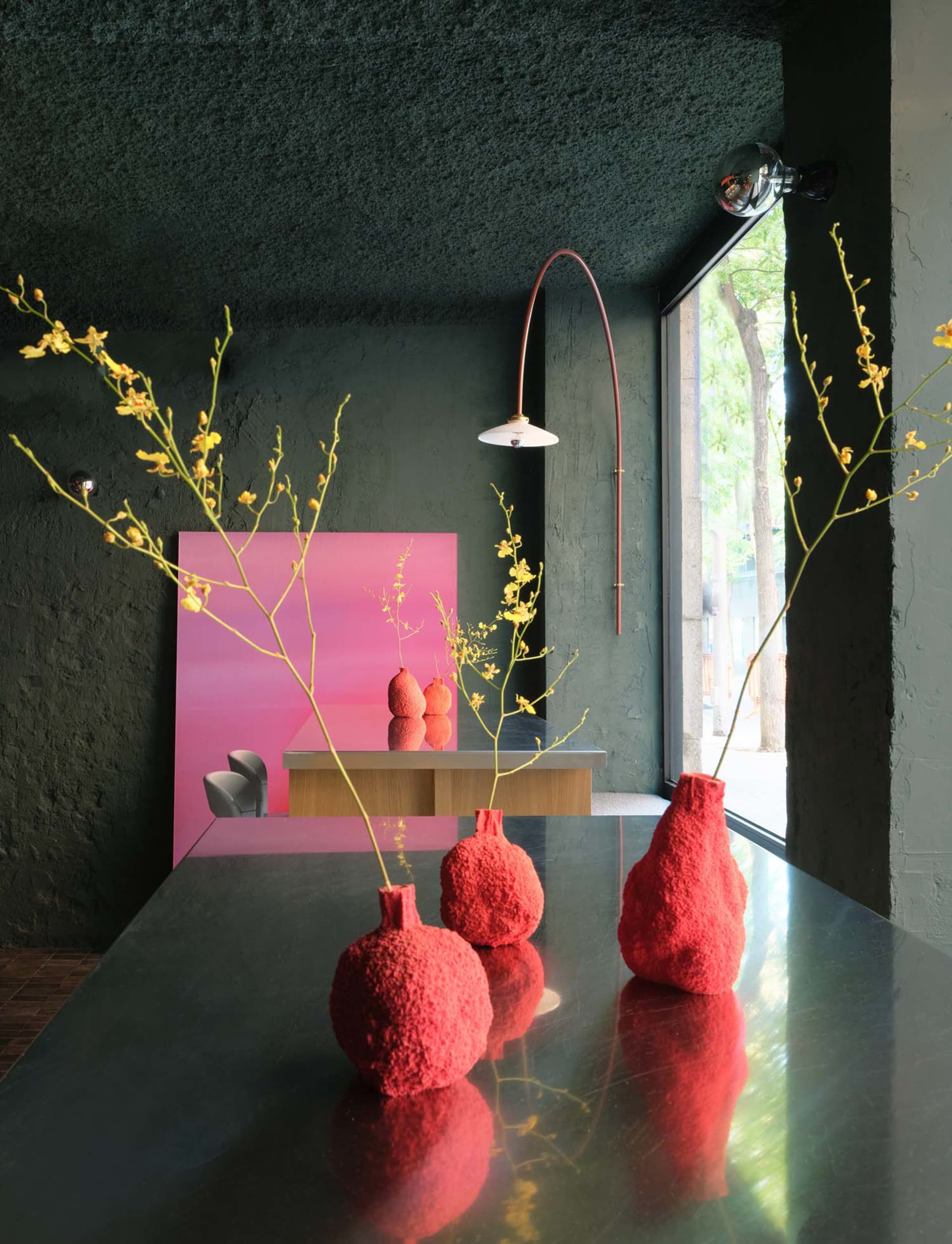A modern living area with a dining table, red vases, and flowers, surrounded by chairs and a potted plant, with a warm and inviting ambiance