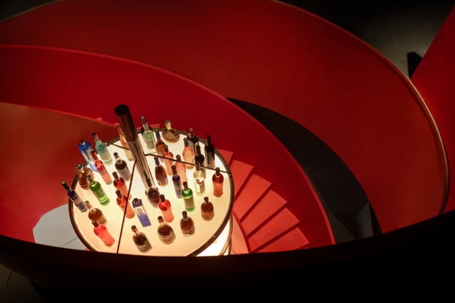 A spiral staircase with a red wall and a large number of bottles displayed in various positions, creating a lively and bustling environment