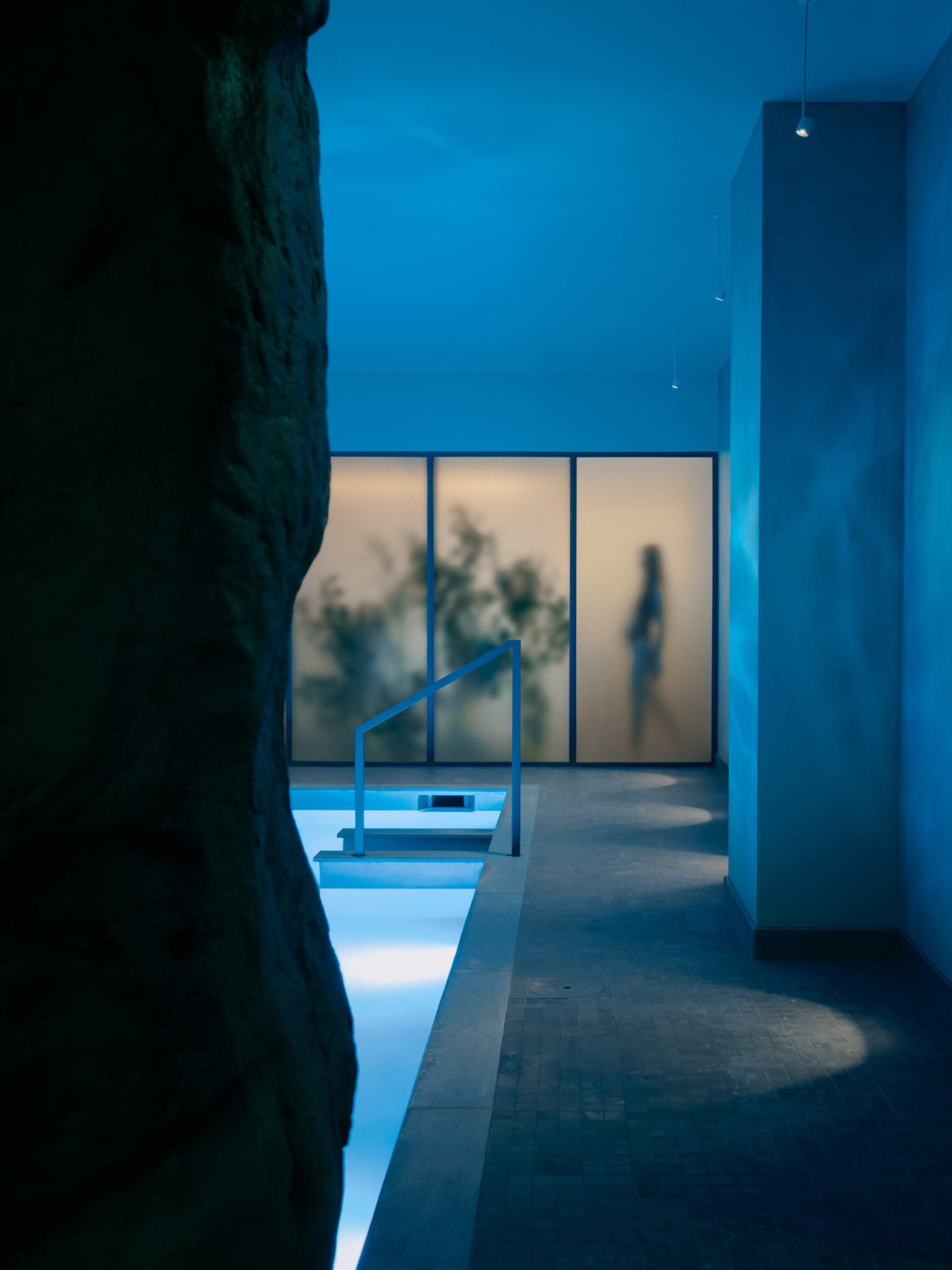 A modern indoor pool area with a blue-lit atmosphere, surrounded by a blue wall and a large rock, creating a relaxing and visually striking environment