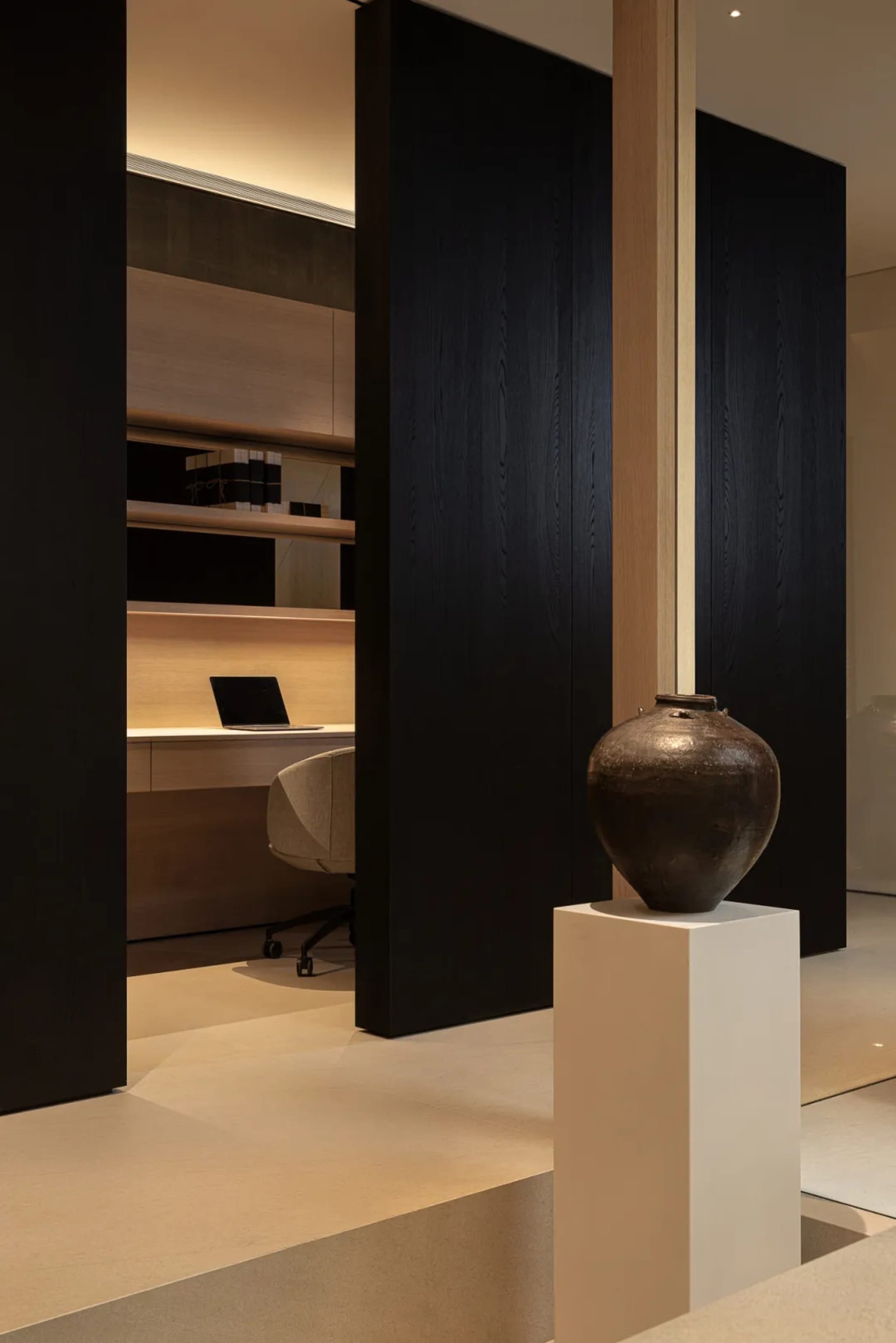 A modern office space with a wooden floor, black walls, a desk, chair, and a vase on a pedestal