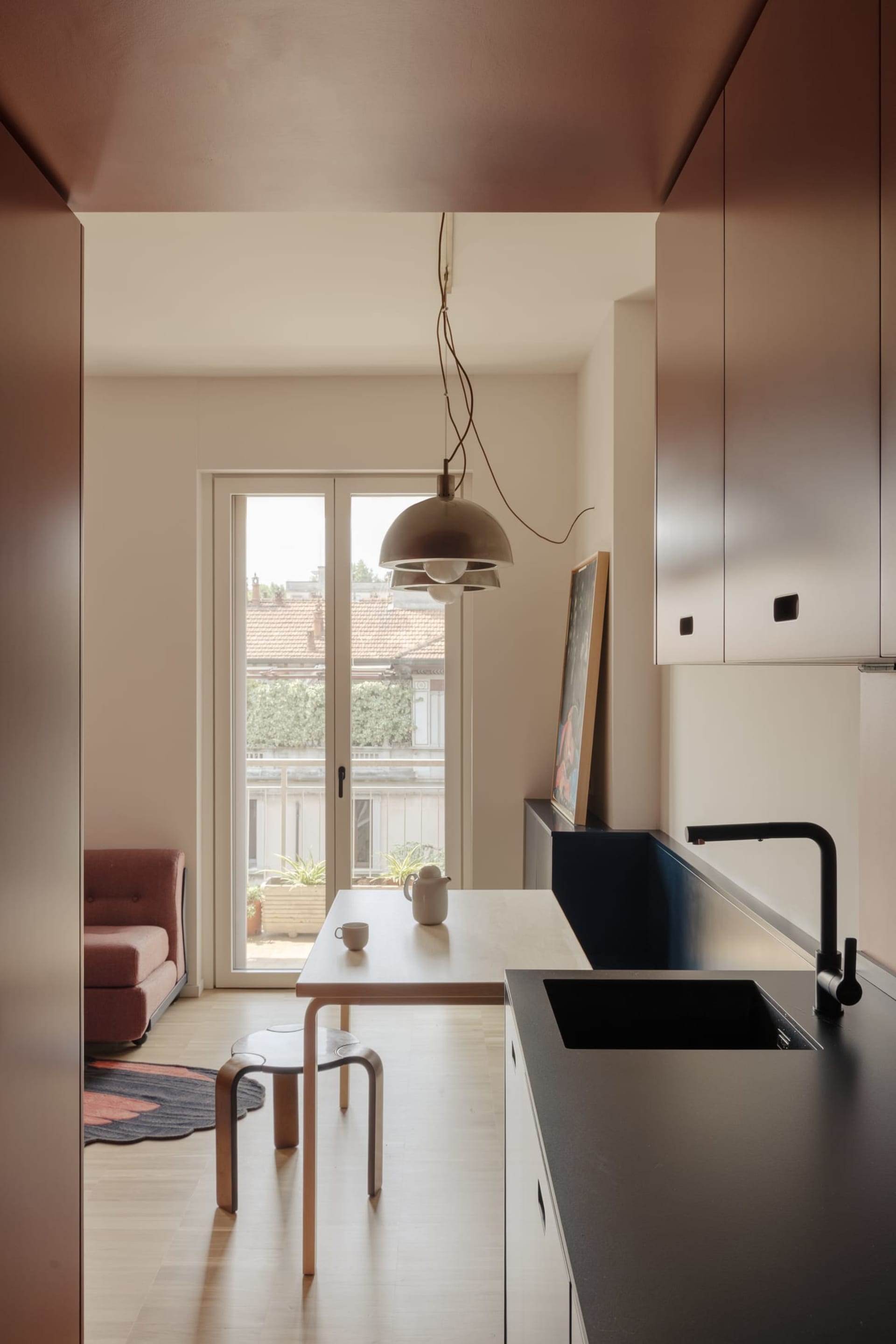 A modern kitchen and living room area with a sink, dining table, couch, and chair, decorated with a potted plant and vase, with natural light streaming in through the windows