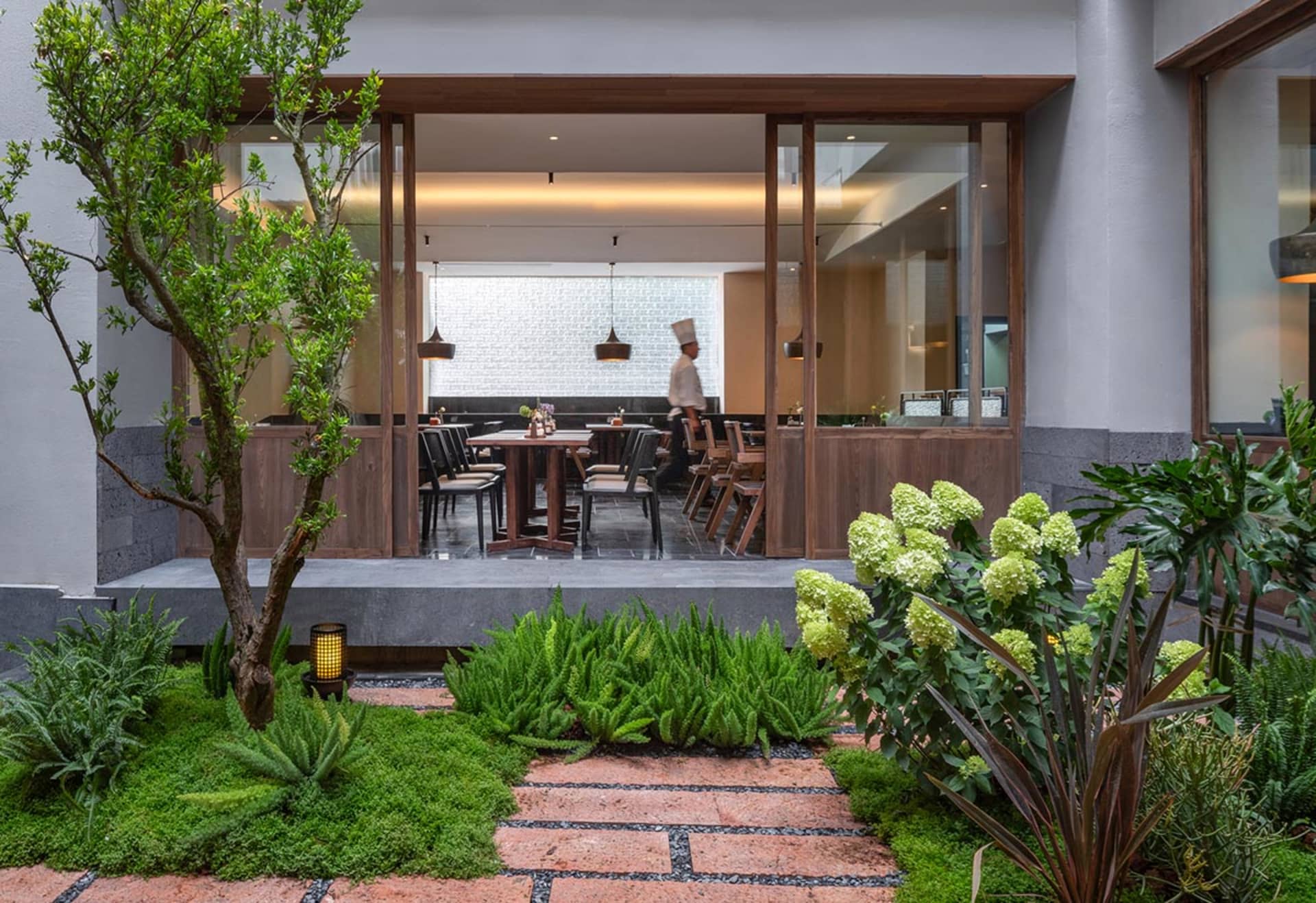 A modern restaurant with a dining area featuring a wooden table and chairs, and a kitchen with metal appliances and a neutral color scheme