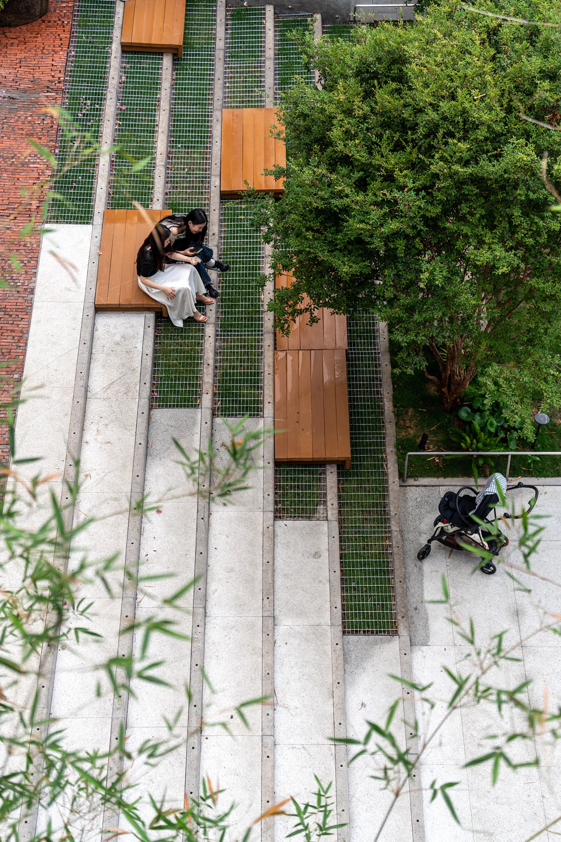 A woman sits on a wooden bench near a tree in a park, with another person standing nearby, creating a peaceful outdoor scene