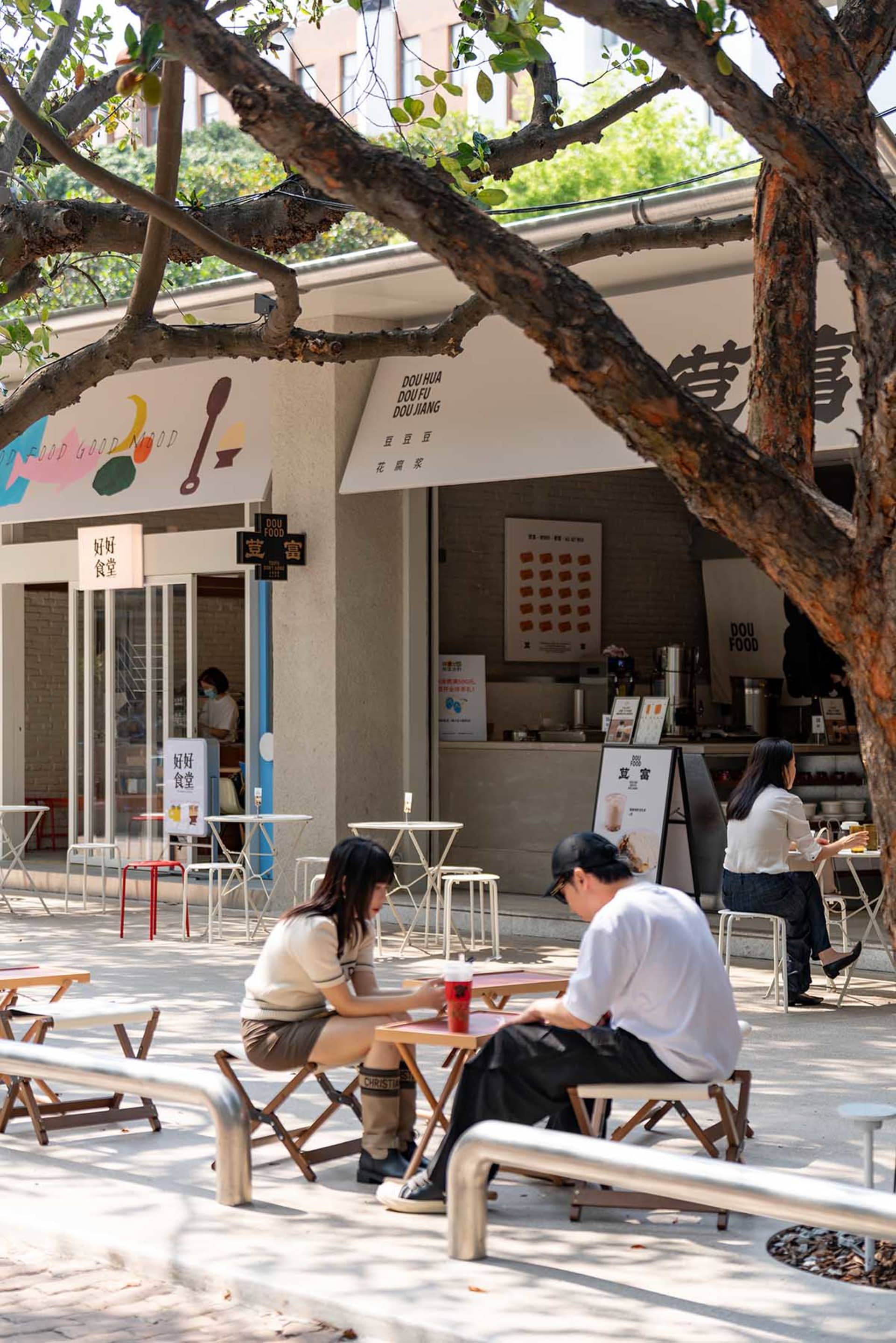 A casual outdoor dining area with people sitting at tables and chairs under a tree, surrounded by a modern restaurant with wood and metal materials, lit by natural sunlight