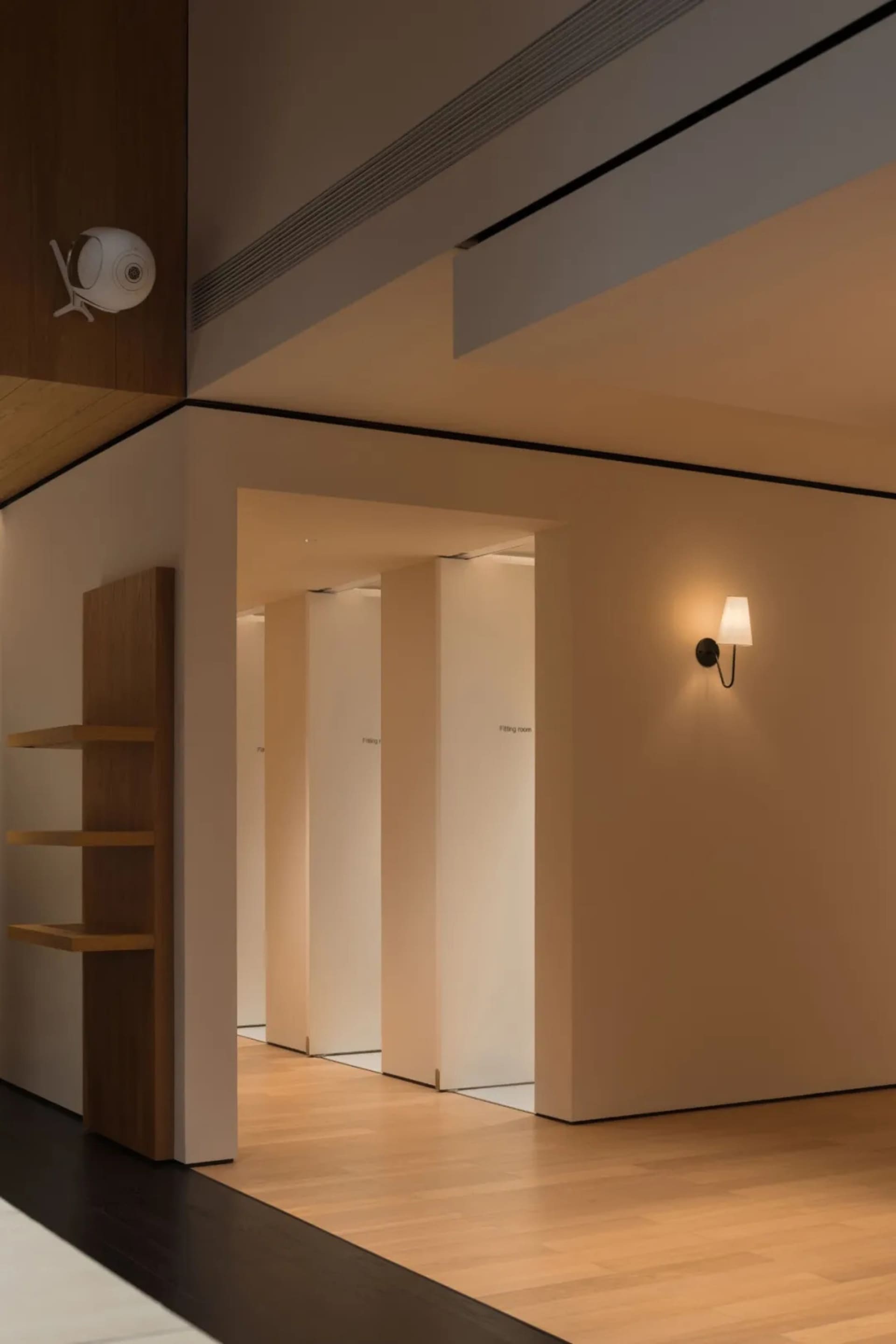 A dimly lit wooden hallway with three doors, each with a light above, in a white color scheme