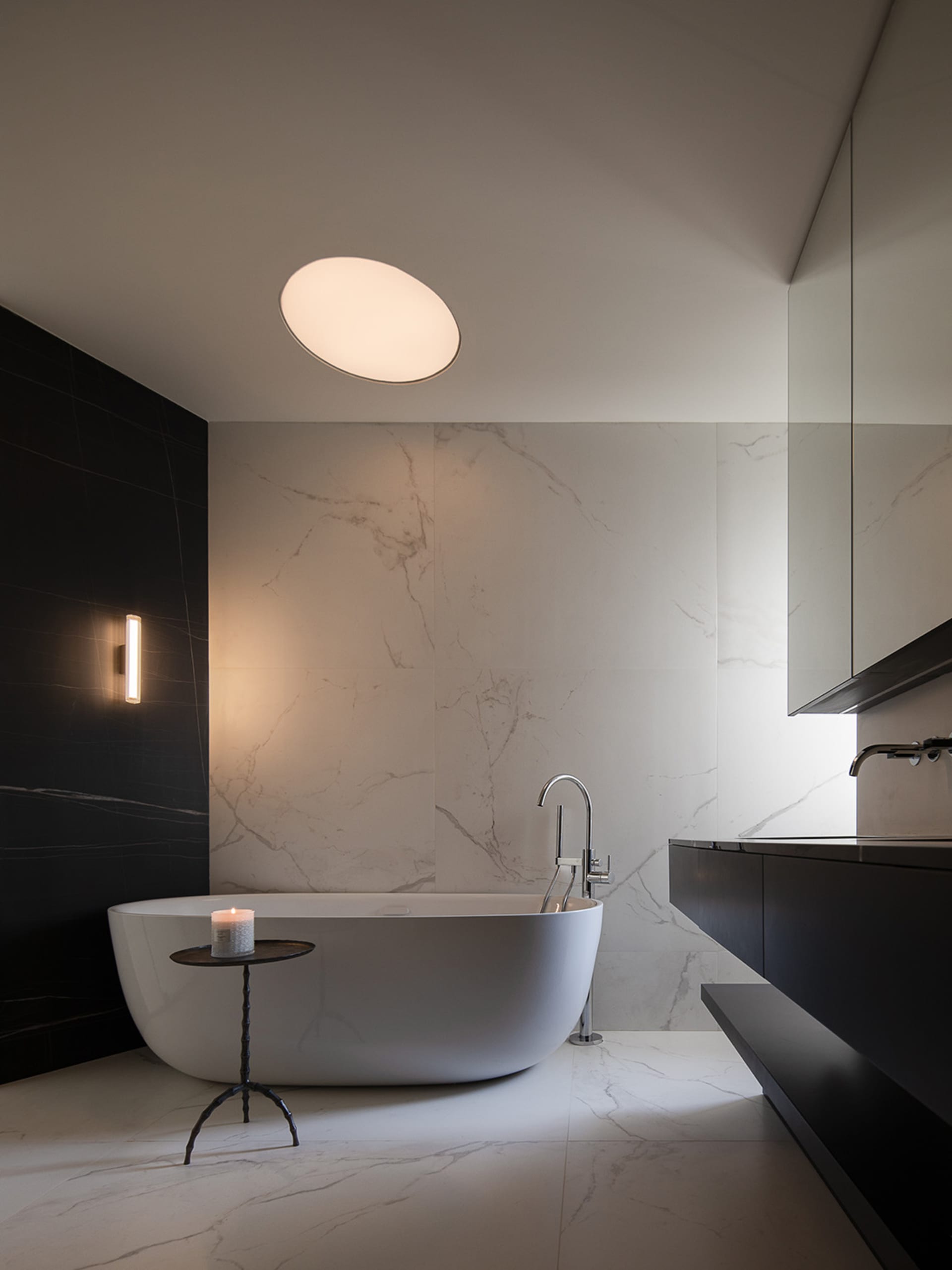 A modern bathroom with a white bathtub, sink, and mirror, featuring a round light fixture and a clean, minimalist aesthetic