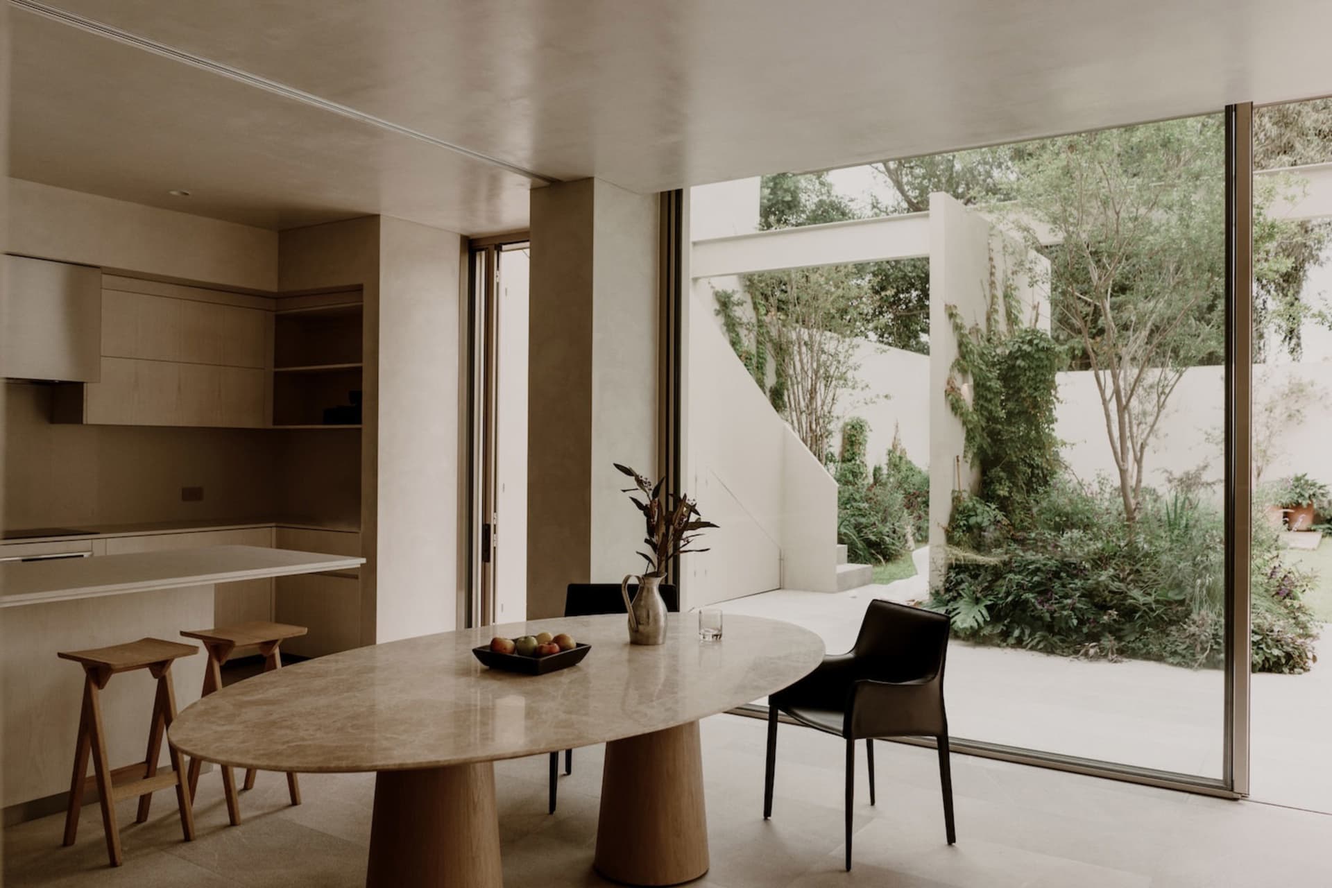 A modern living room with a dining table, chairs, and a potted plant, surrounded by a large window allowing natural light to enter the space