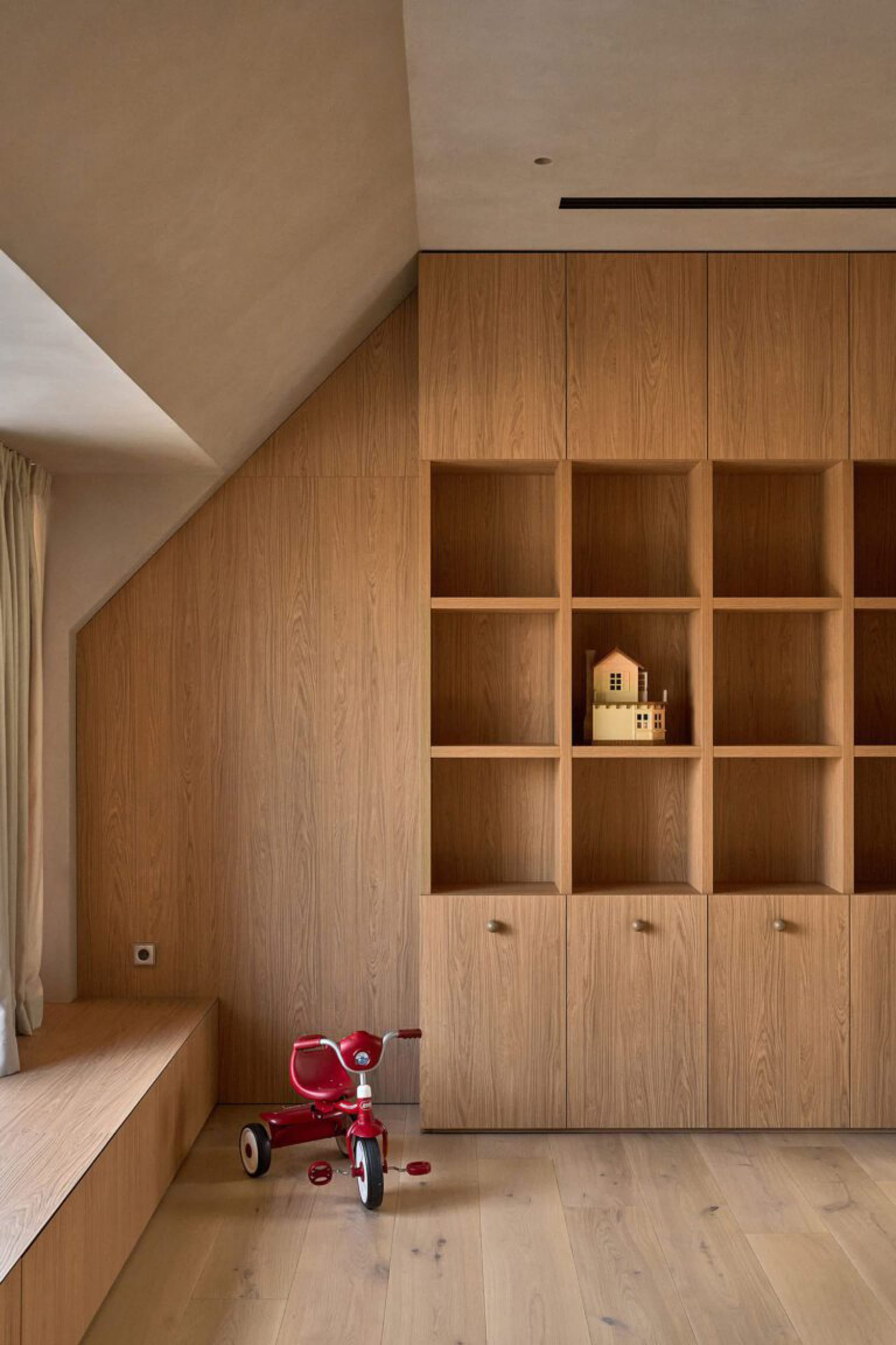 A dimly lit room with wooden walls and floor, featuring a wooden bookshelf with a toy house and a red toy car, creating a cozy and intimate atmosphere