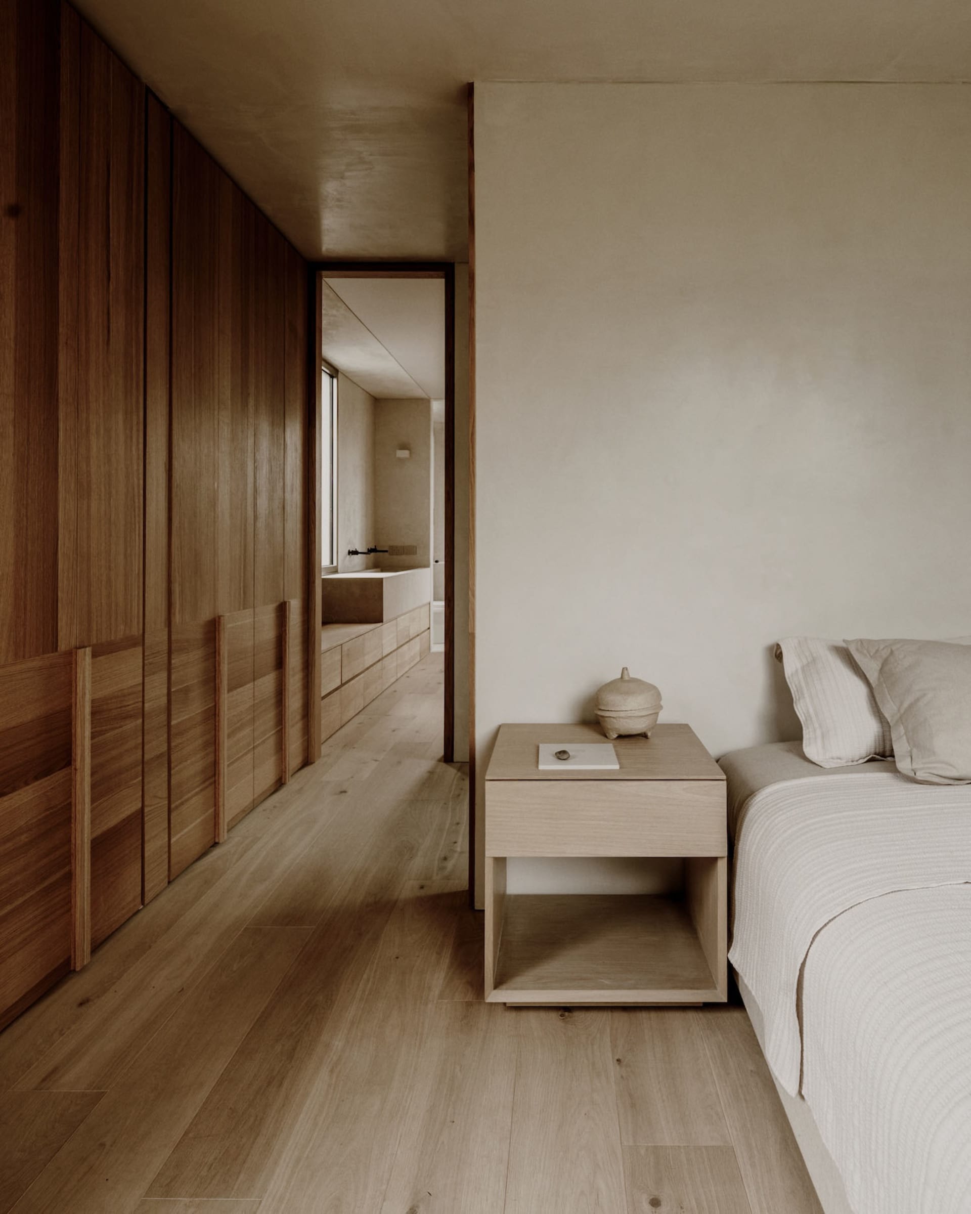 A modern bedroom with a wooden floor, a bed on the right, a table in the middle, and a mirror above, lit by natural sunlight through a window