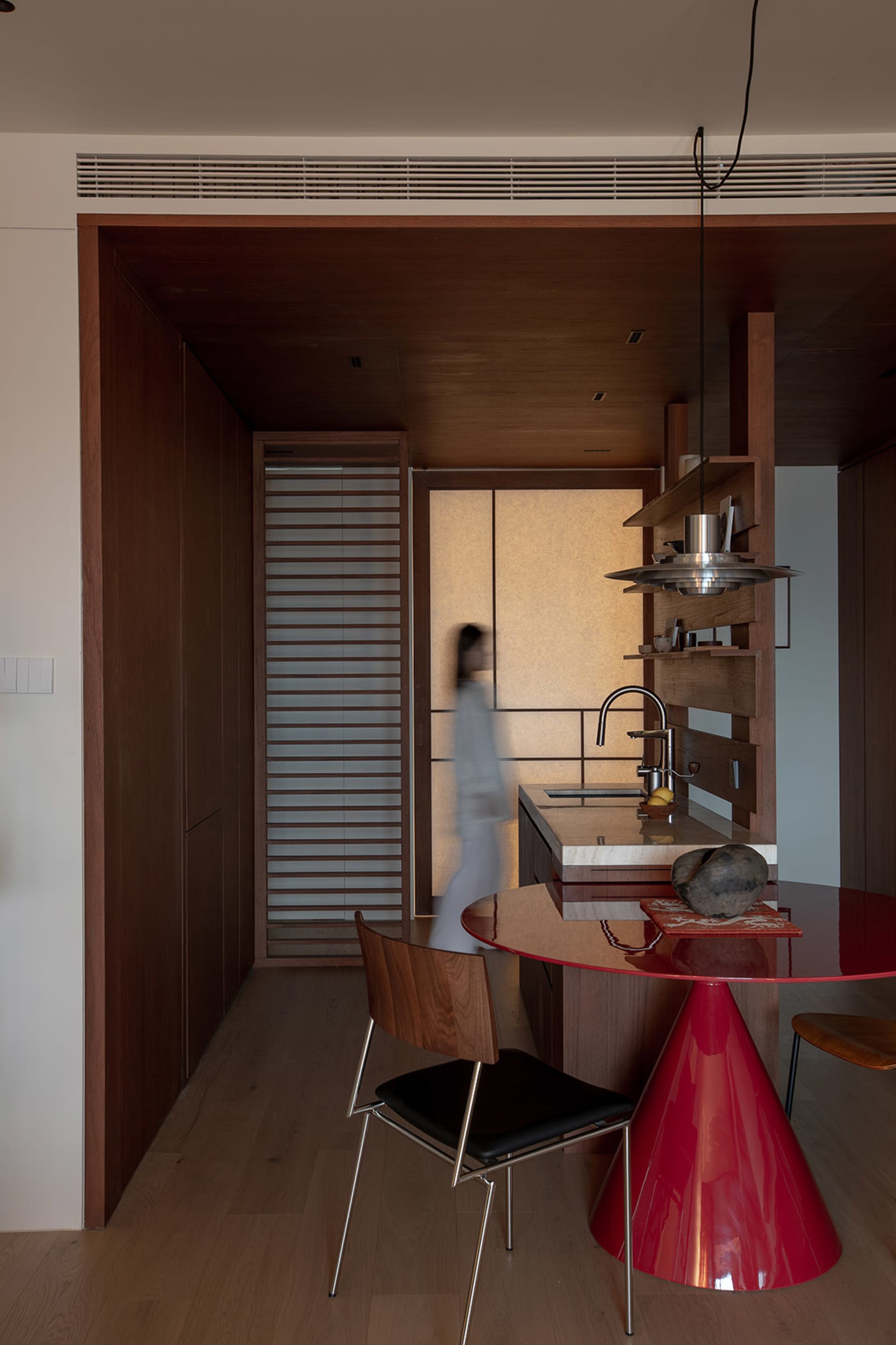 A modern kitchen with a wooden floor, a red dining table, and a variety of furniture and items, lit by natural sunlight
