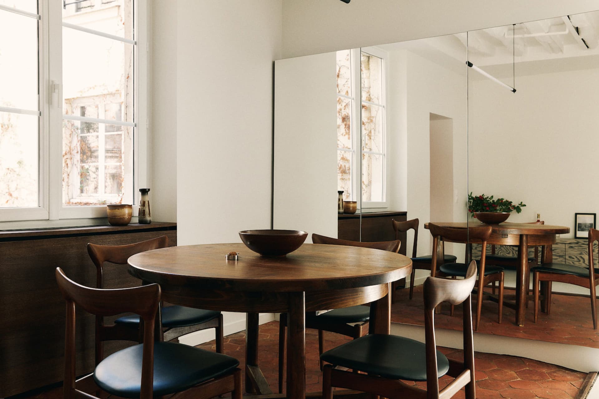 Dining room with a wooden table and chairs, surrounded by a wooden floor and a mirror, with natural light and a warm atmosphere