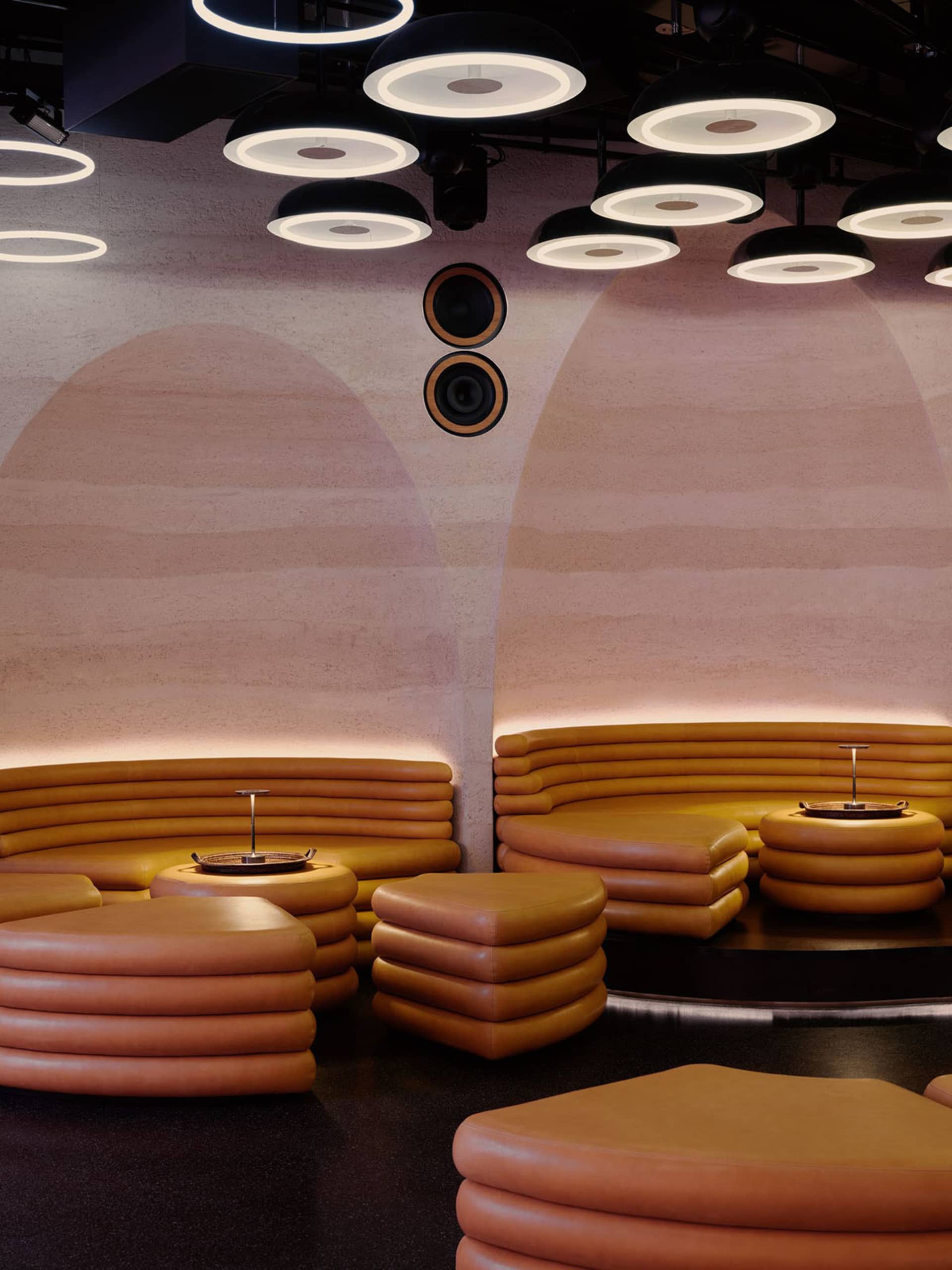 A modern lounge with a circular layout, featuring a central table surrounded by semi-circular couches, illuminated by natural and artificial light, creating a warm and inviting atmosphere