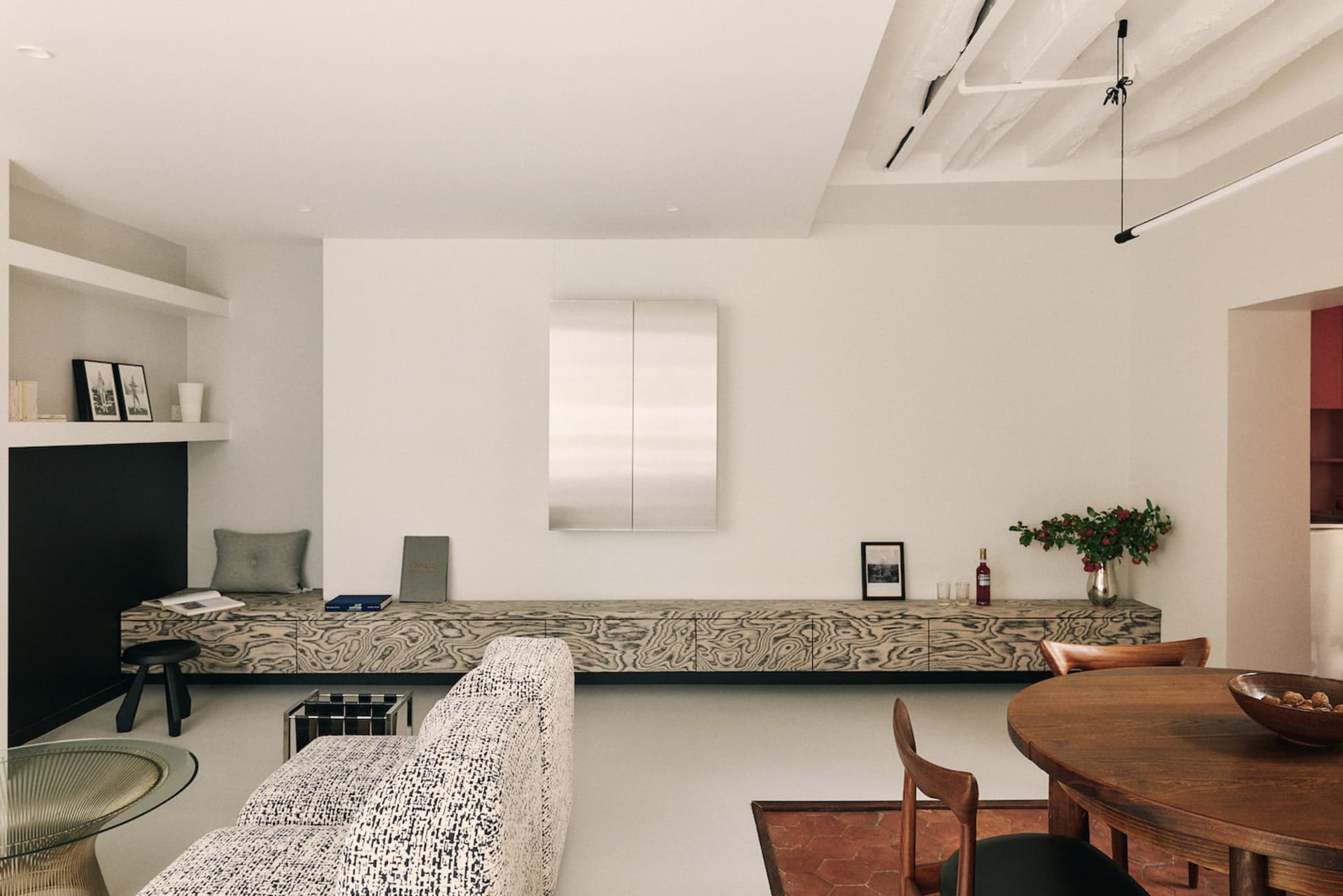 A modern living room with a wooden dining table and chairs, a couch, a chair, and a refrigerator, well-lit with natural light from the windows