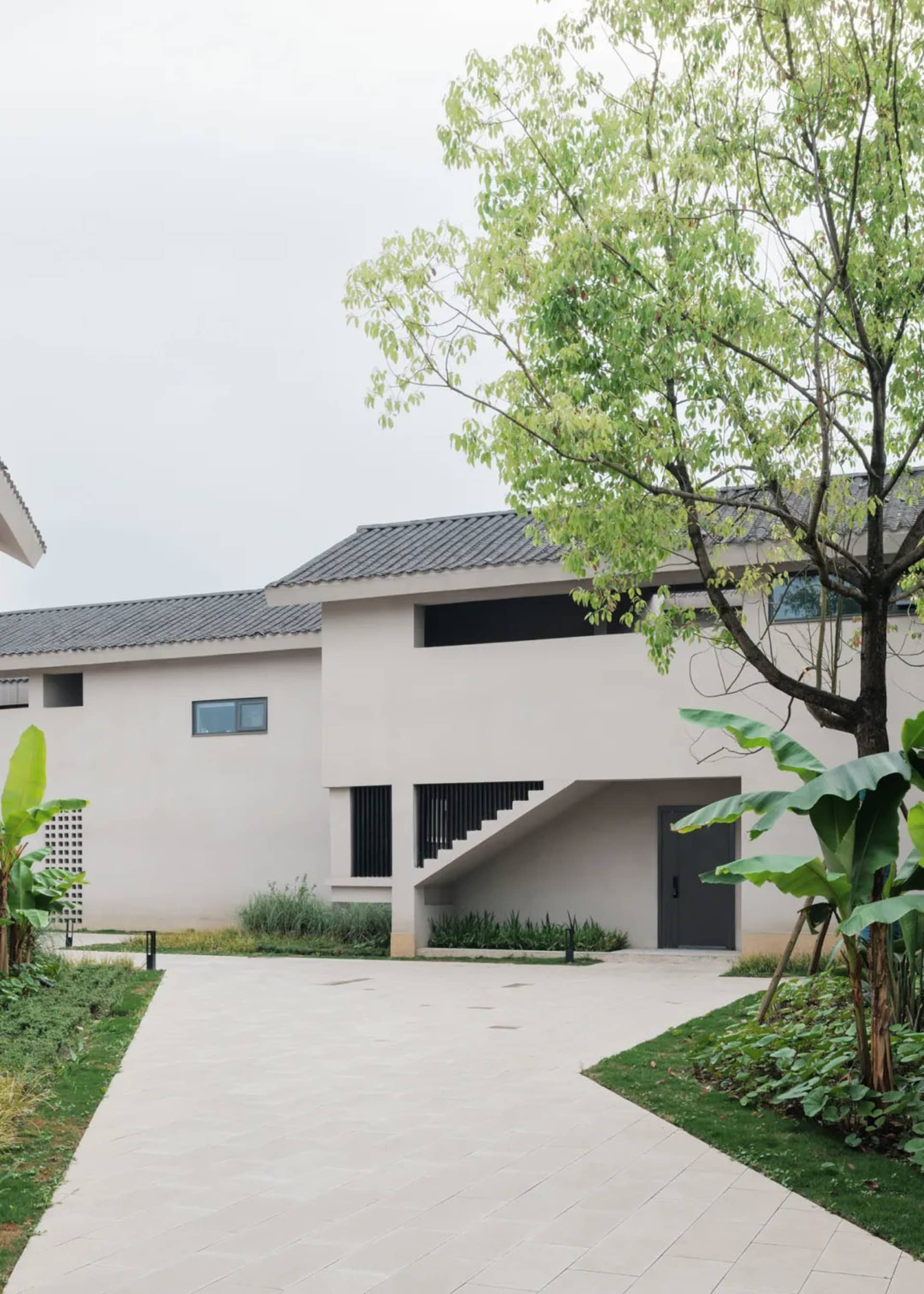 A modern house with a white exterior, large window, and staircase, set in a residential area with a tree in the background, under natural sunlight