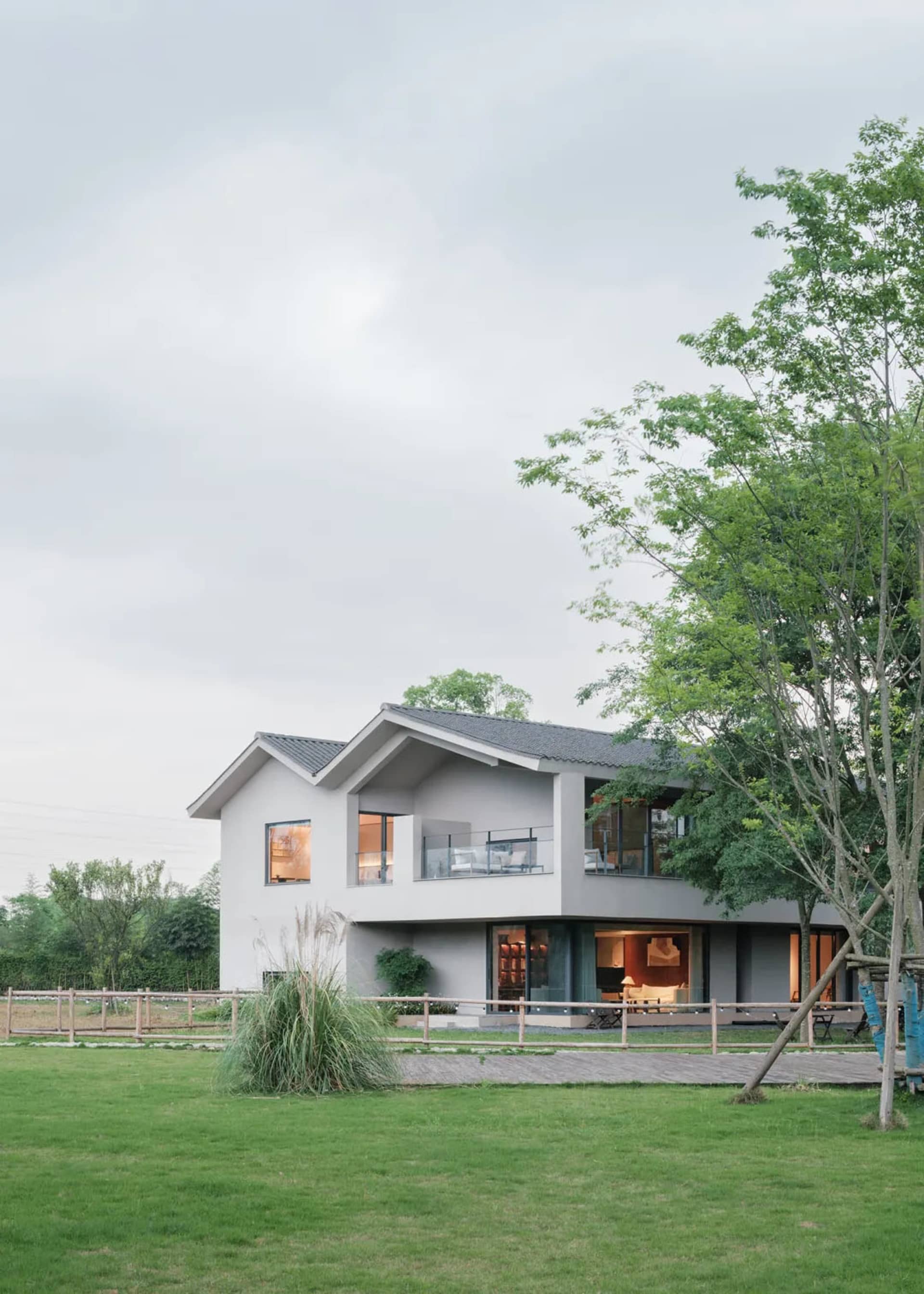 A modern house with a large white exterior, surrounded by a lush green field, featuring a deck and patio area with a bench and dining table, and a well-lit interior with comfortable furniture