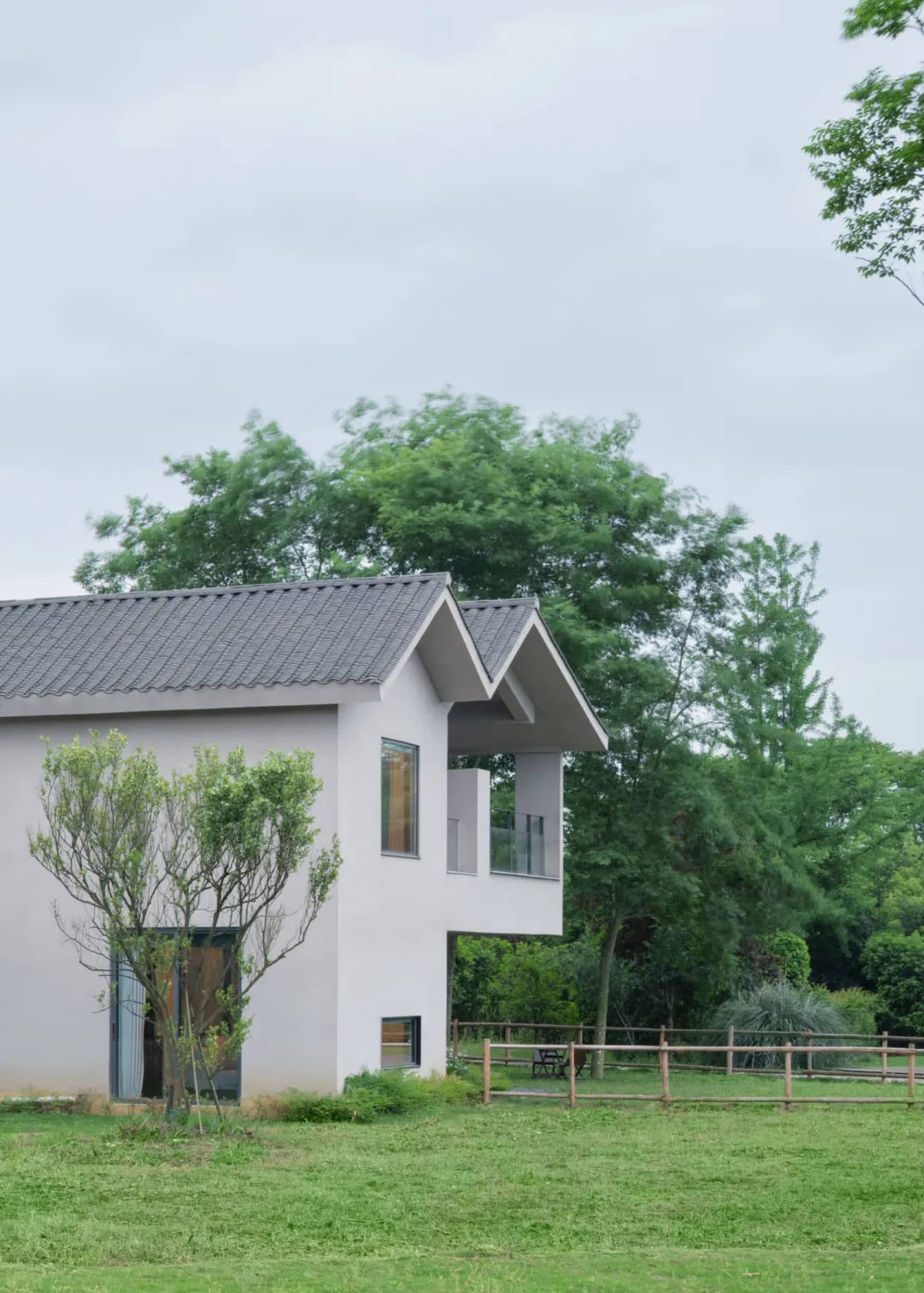 A modern house with a white exterior and gray roof, surrounded by trees, with a large window allowing natural light to enter the space