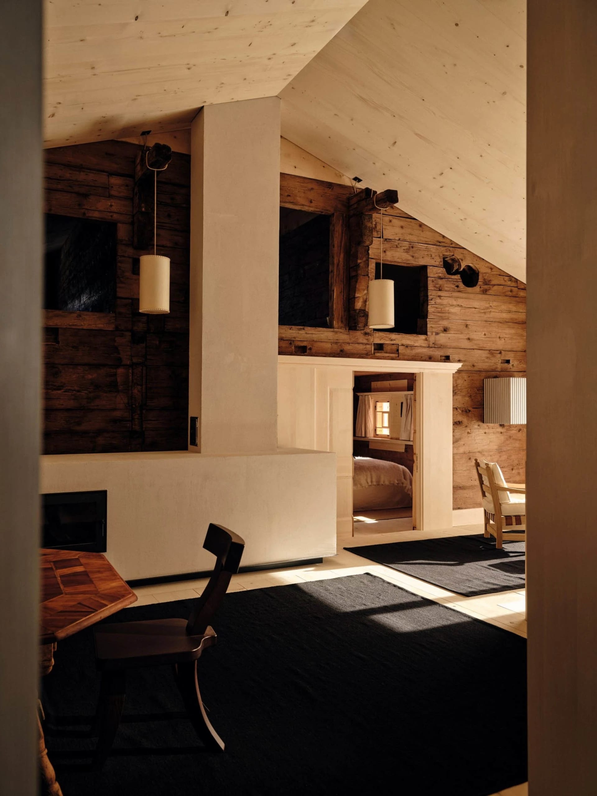 A well-lit living room with a wooden floor, a fireplace, a dining table, a couch, a chair, and a bed, giving a cozy and rustic feel
