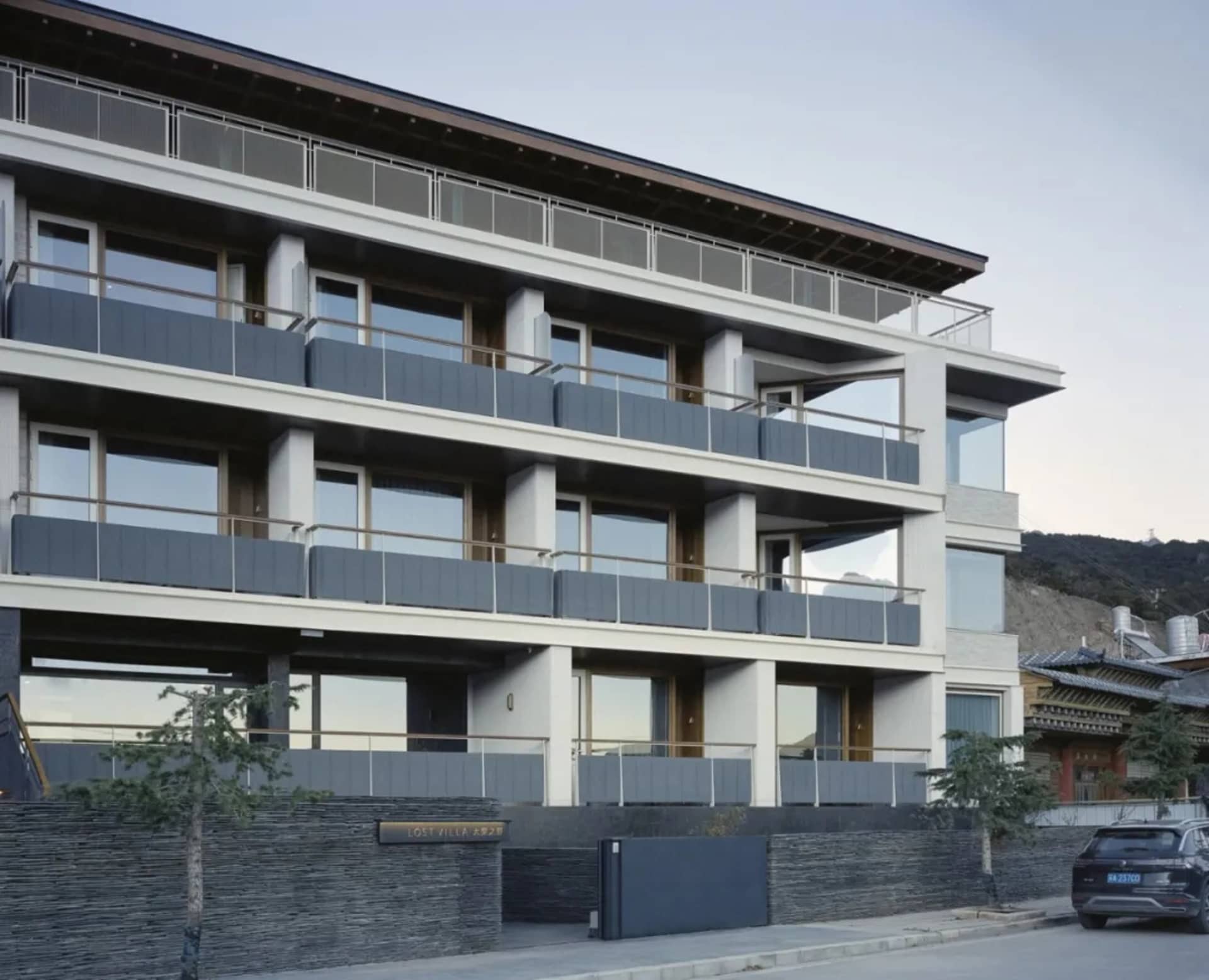 Modern multi-story building with a balcony, black railing, and a backdrop of mountains