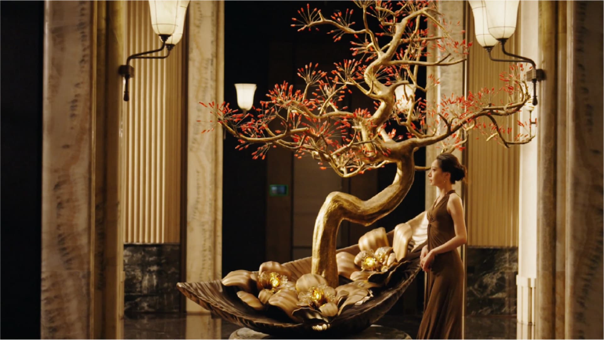 A woman stands in a dimly lit living area with a gold tree sculpture, a couch, and a chair, surrounded by modern decor with gold and brown tones