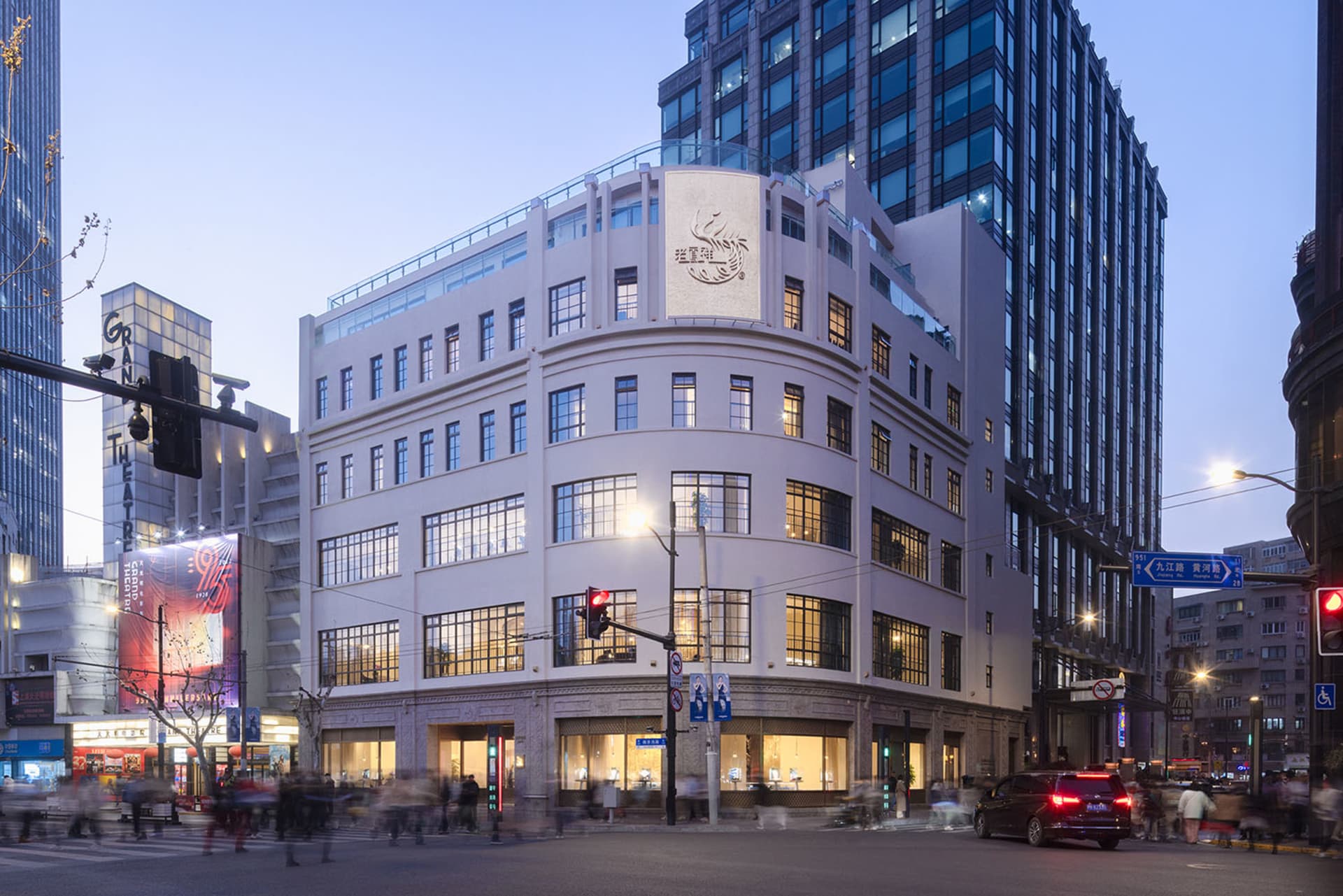 A large modern building with many windows, set against a busy urban backdrop with people walking and traffic lights visible