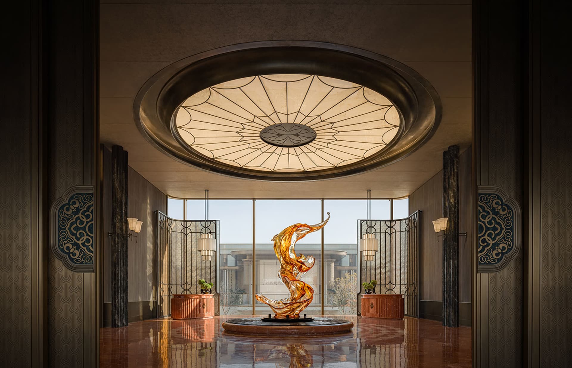 Bright and inviting lobby with a glass ceiling, a circular light fixture, a large sculpture, and a fountain, illuminated by natural light