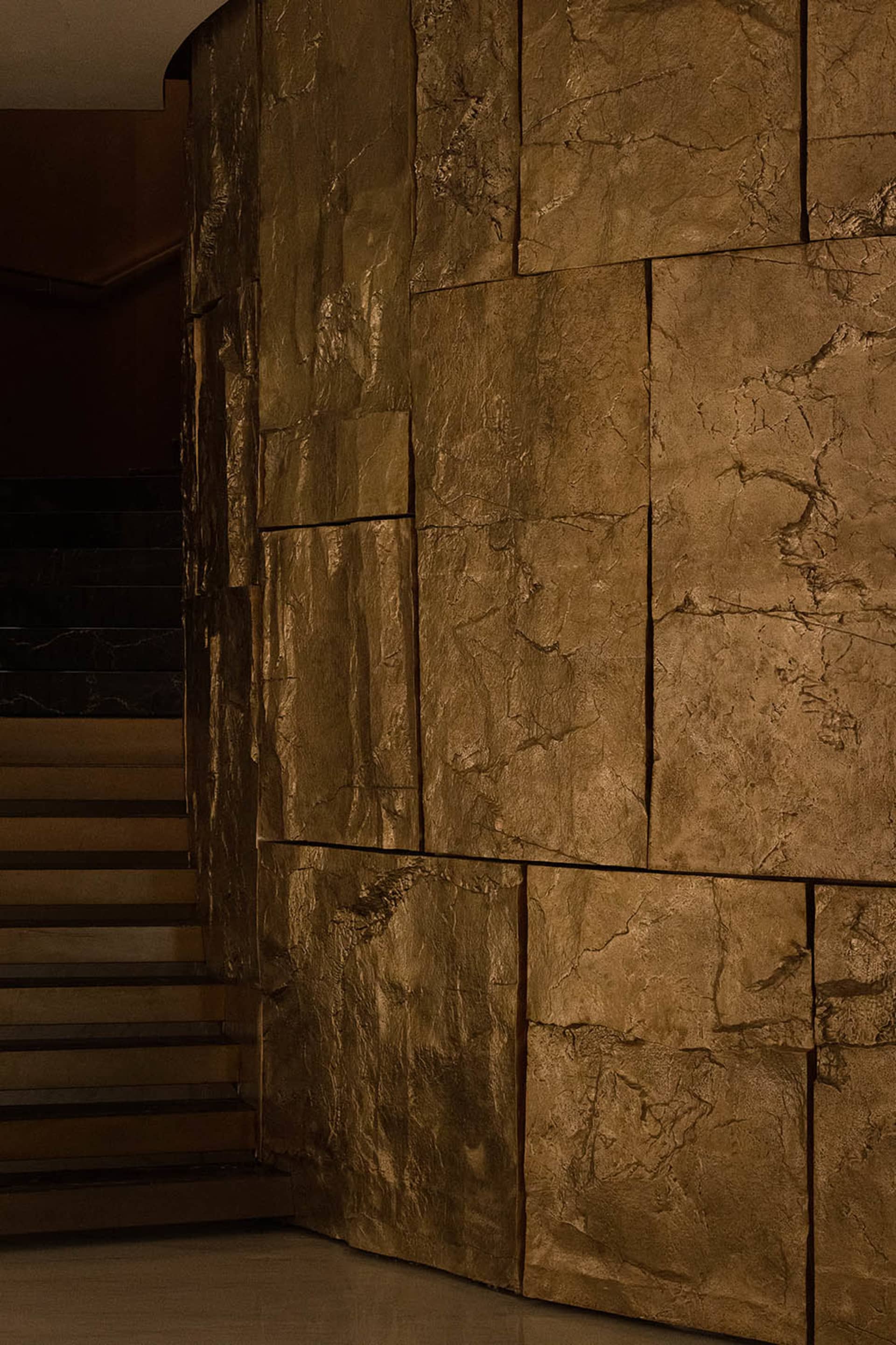 A stone wall with a textured surface and a concrete staircase with a metal railing in the background, set in a dimly lit outdoor area