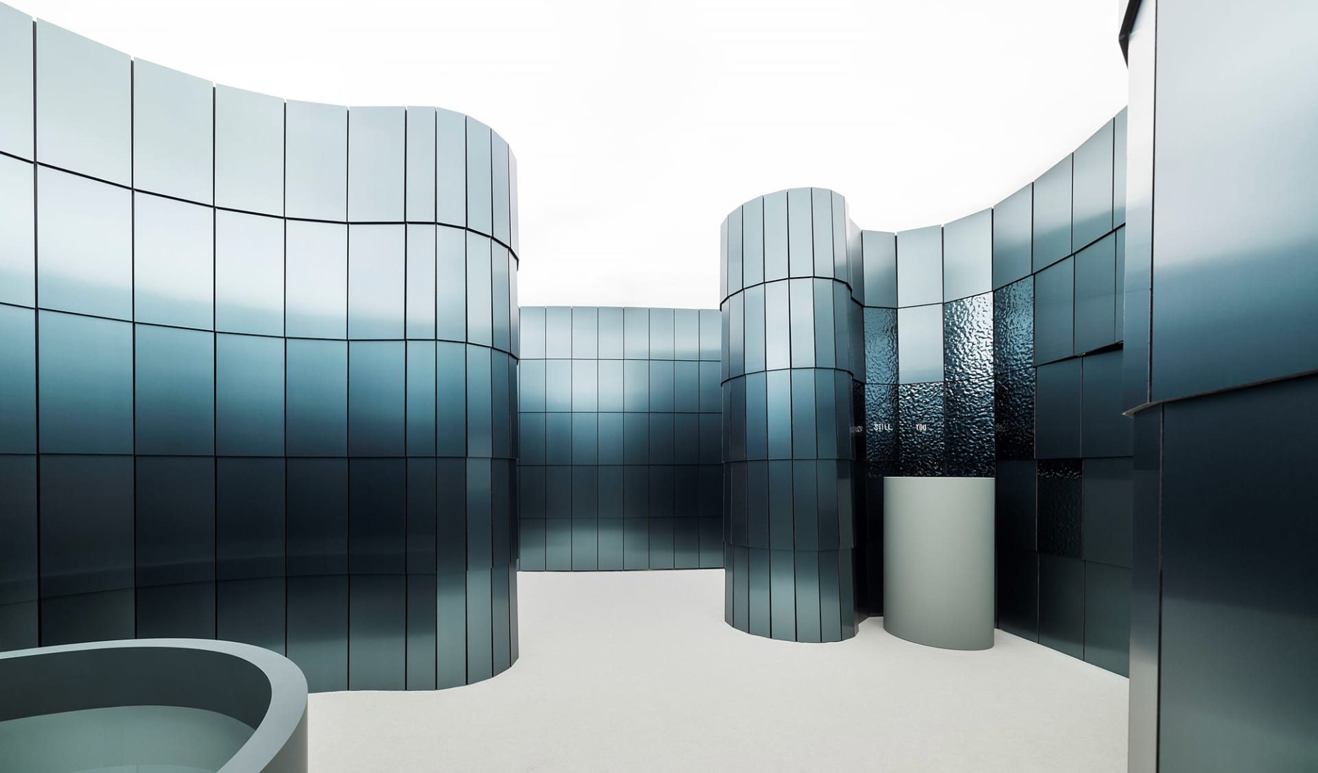 A modern building with a large glass wall reflecting the sky, surrounded by metal and glass materials, with natural light and a bright atmosphere