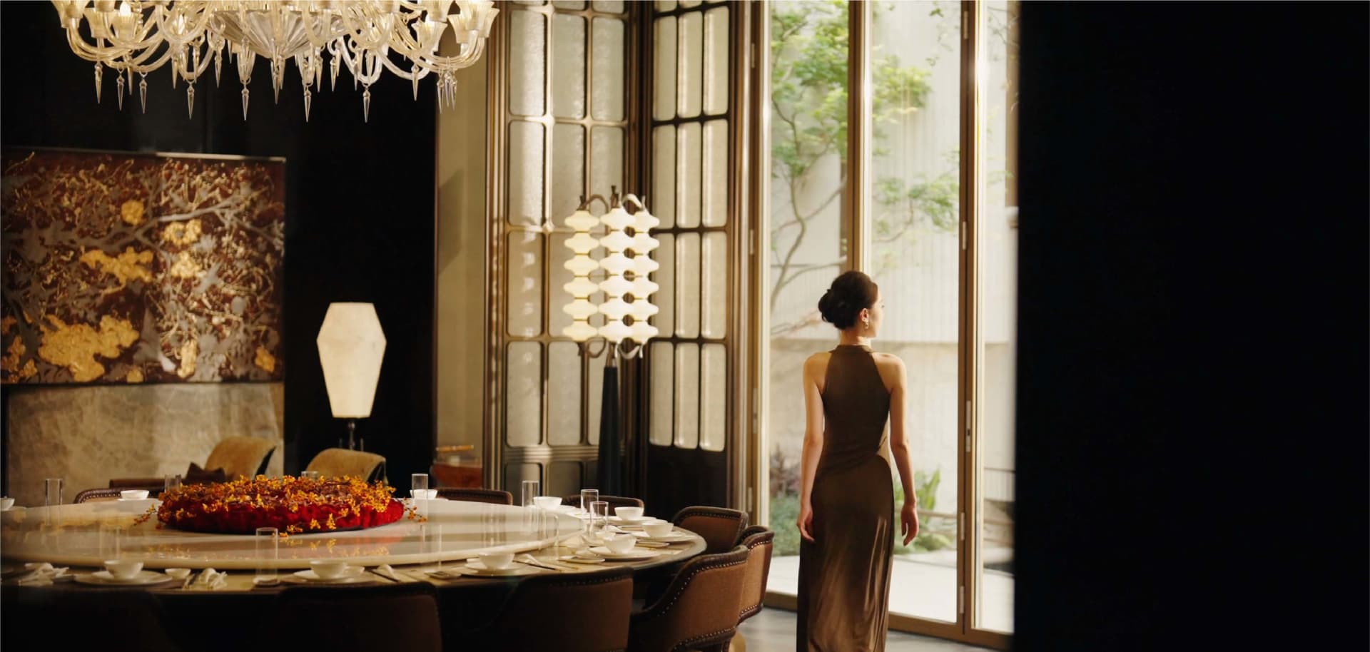 A woman stands in front of a window in a large, elegant dining room with a wooden dining table and chairs, surrounded by wine glasses