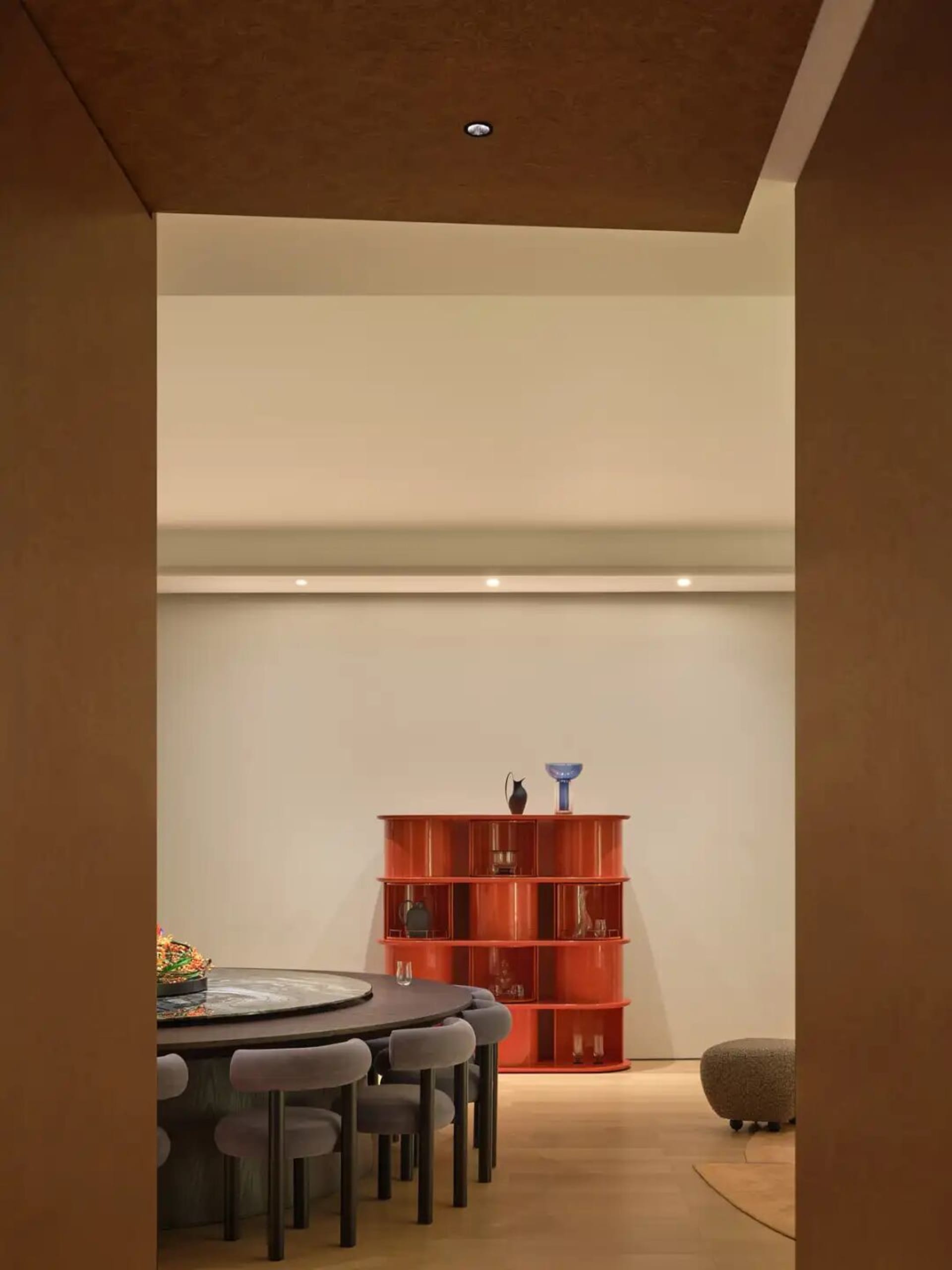 A modern study with a wooden floor, a dining table, a red bookshelf, and a chair, lit by natural sunlight