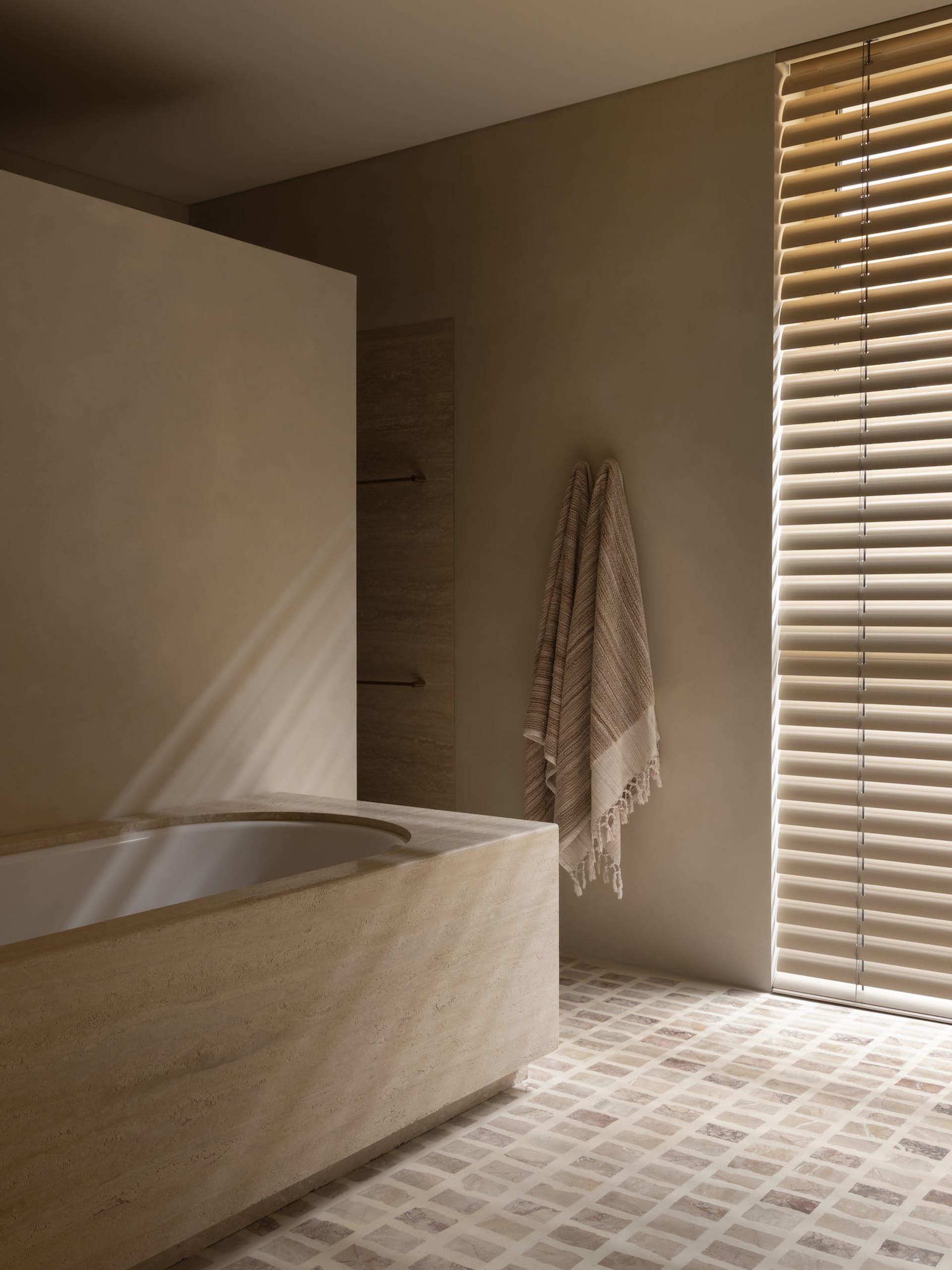 A well-lit bathroom with a beige color scheme, a bathtub, and a towel, featuring natural stone or tile materials and a window
