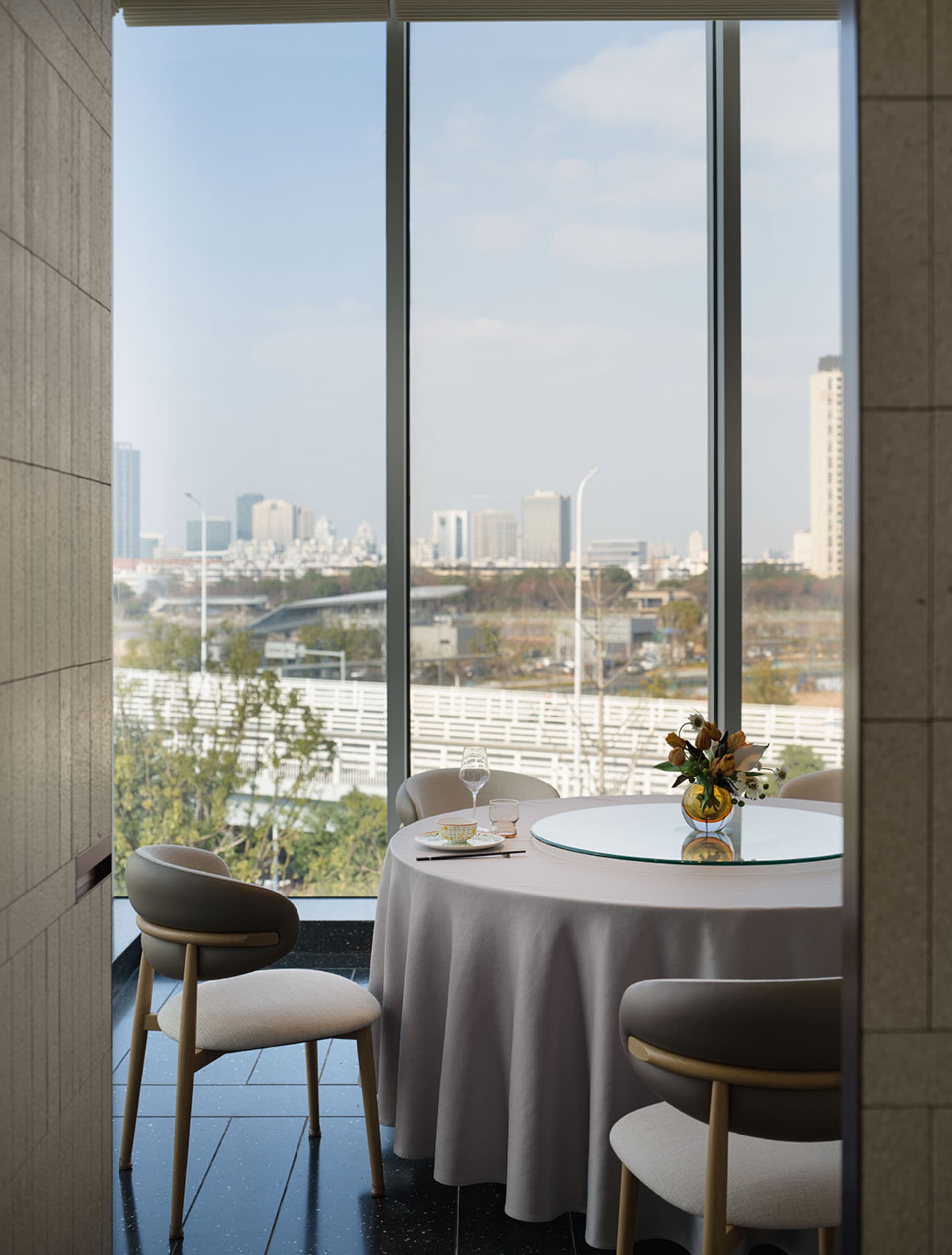 Dining area with a large window offering a city view, featuring a set table, chairs, and a potted plant, with a modern design and natural lighting