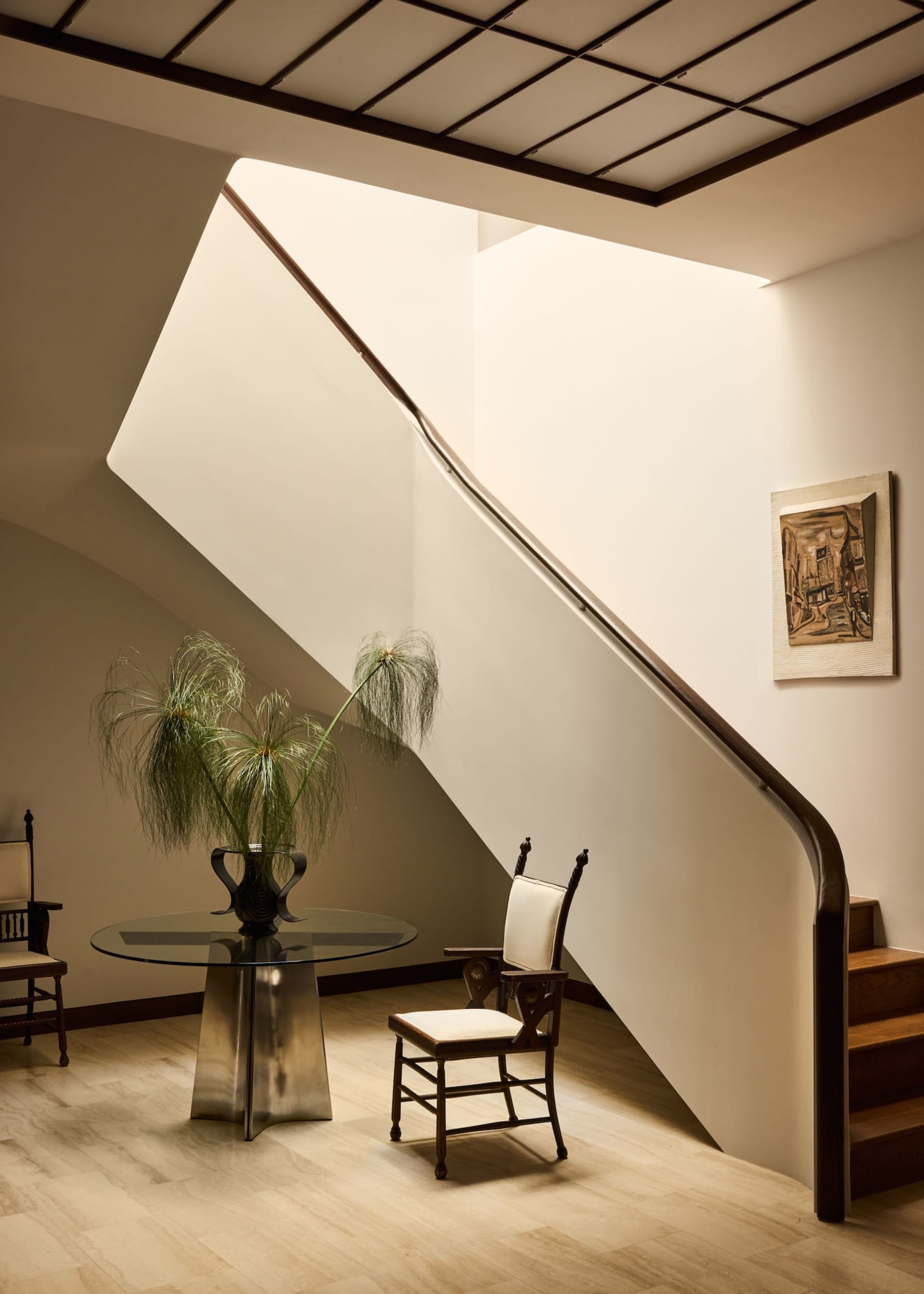 A modern living room with a glass table, wooden staircase, and natural light from windows, featuring a vase and potted plant