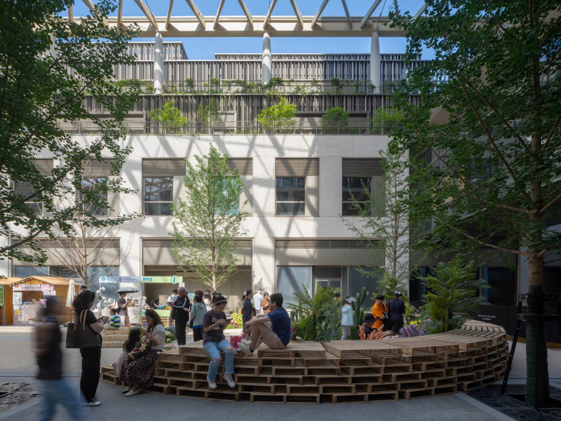 A bustling public space with a wooden bench in the center, surrounded by a modern building with a mix of concrete and wood, and people interacting in the outdoor area