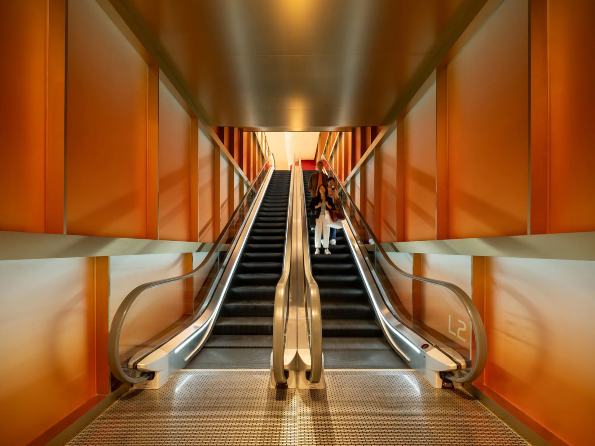Photograph of an orange-colored building with a metal staircase and people on the second floor, taken in bright daytime lighting