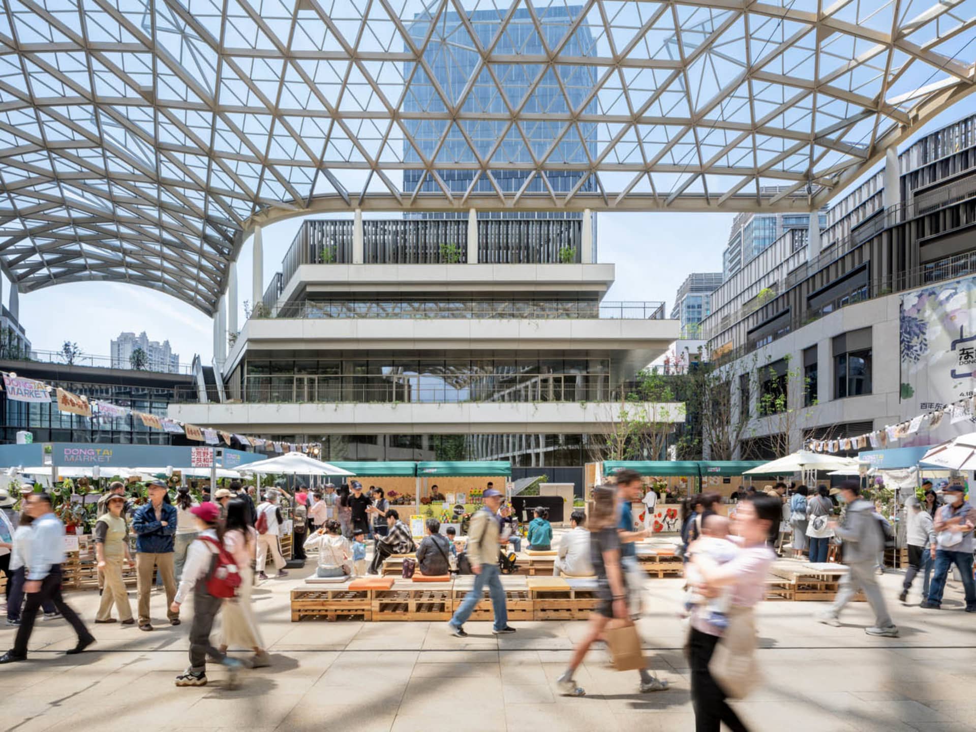 A bustling city street with people walking in various directions, lined with buildings and umbrellas, suggesting outdoor seating or vendors