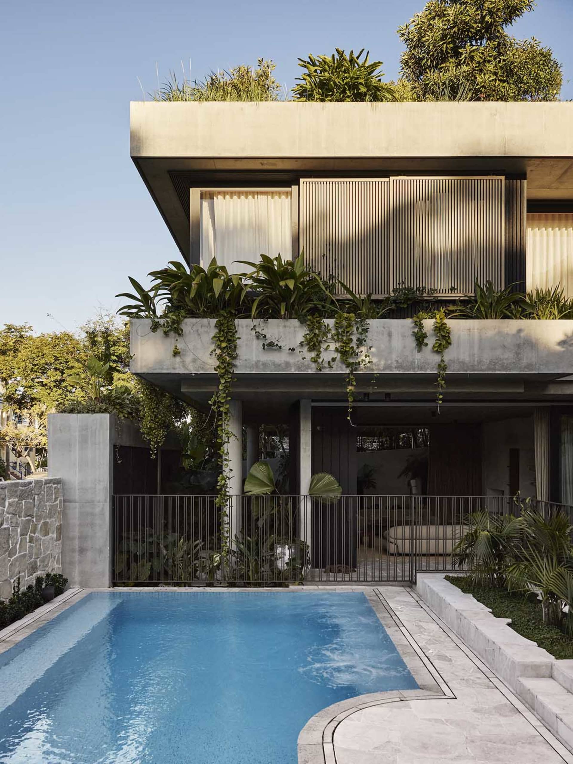 A modern house with a large window, balcony, and blue swimming pool surrounded by a patio and potted plants, with natural light and a clean, minimalist design