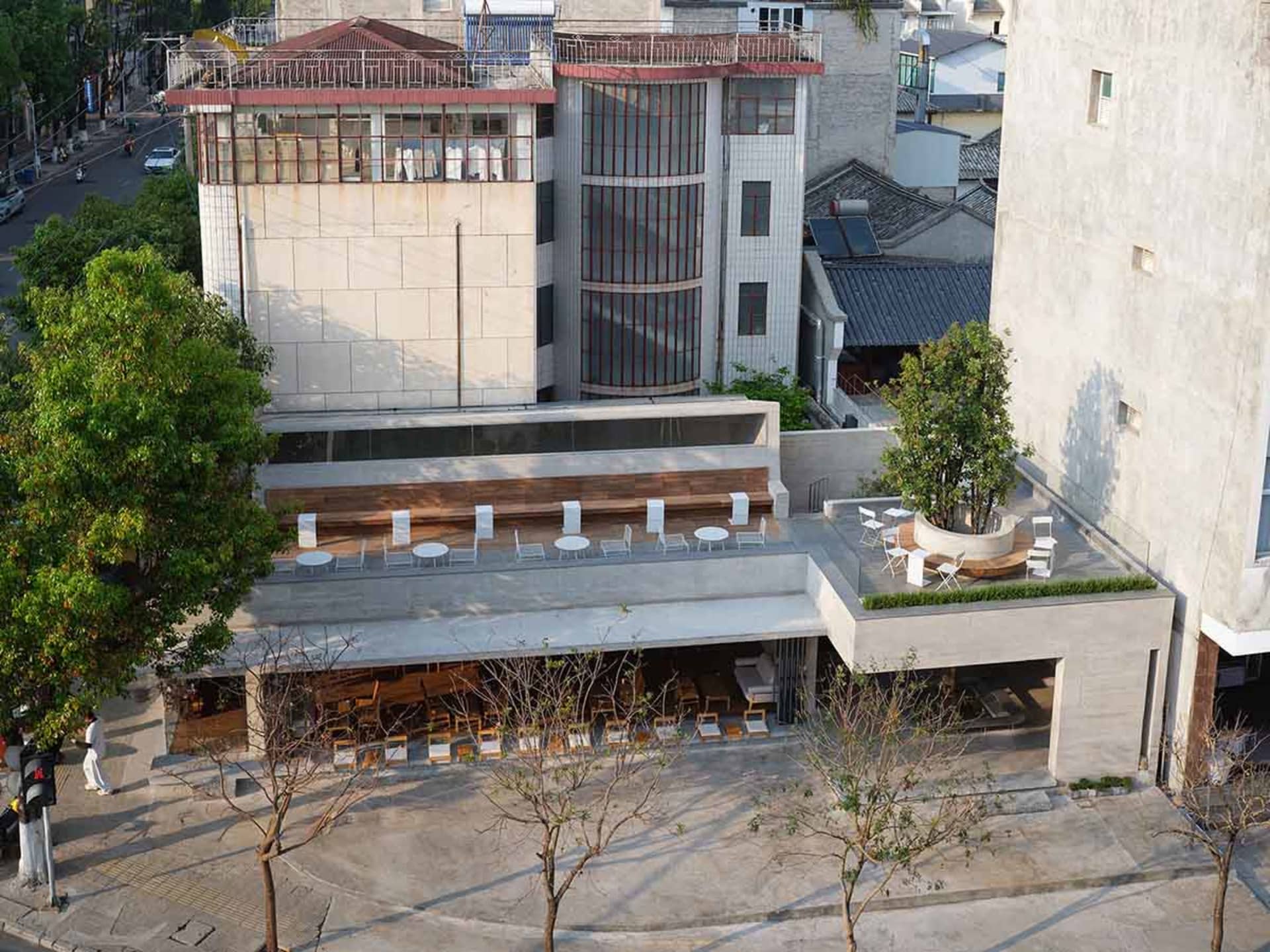 A modern building with a large glass wall and rooftop terrace, featuring a furnished outdoor seating area with chairs and dining tables, set against a concrete facade in a natural daylight setting
