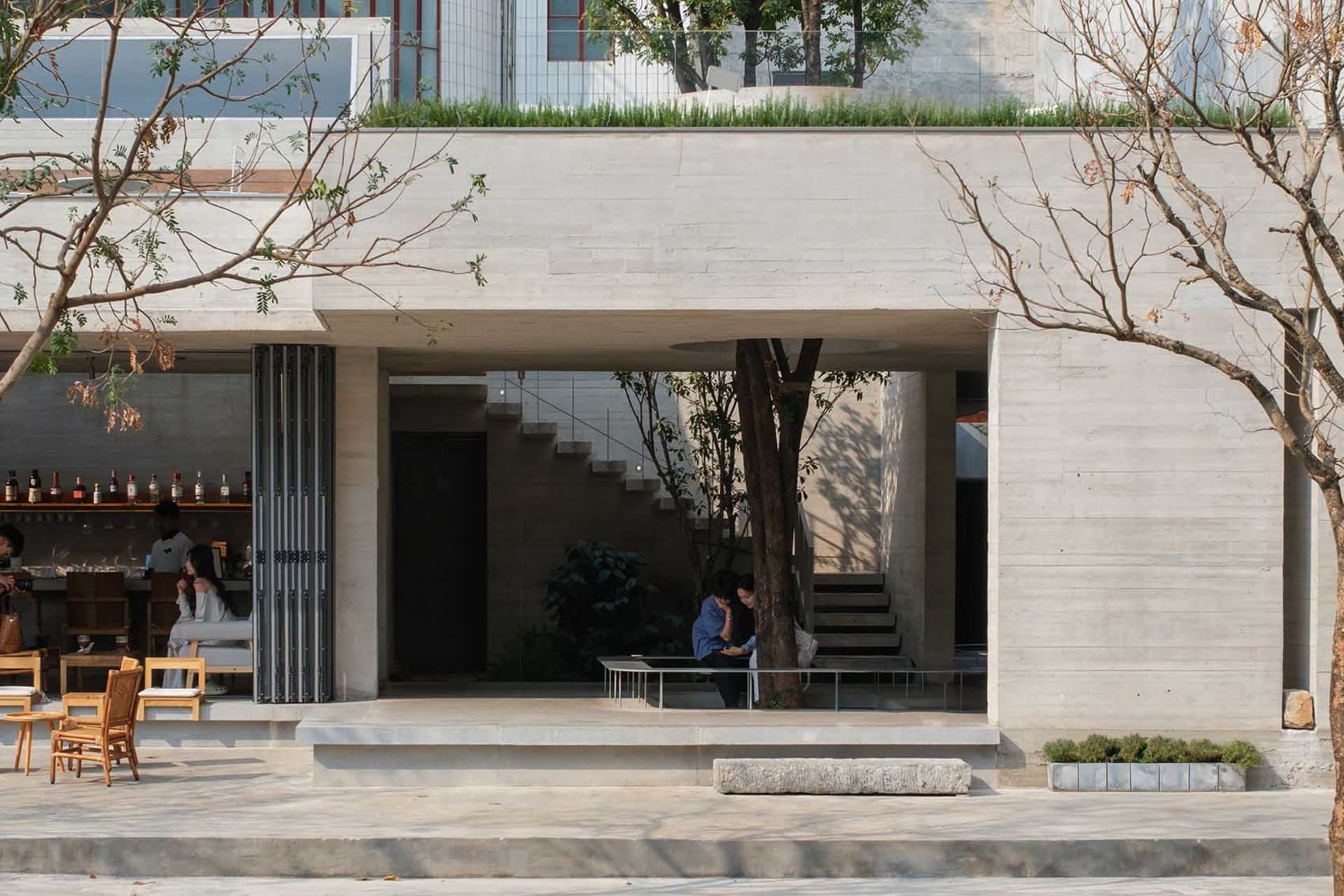 一棟現代住宅，混凝土外觀，庭院設有餐桌椅，前院有樹，極簡當代風格