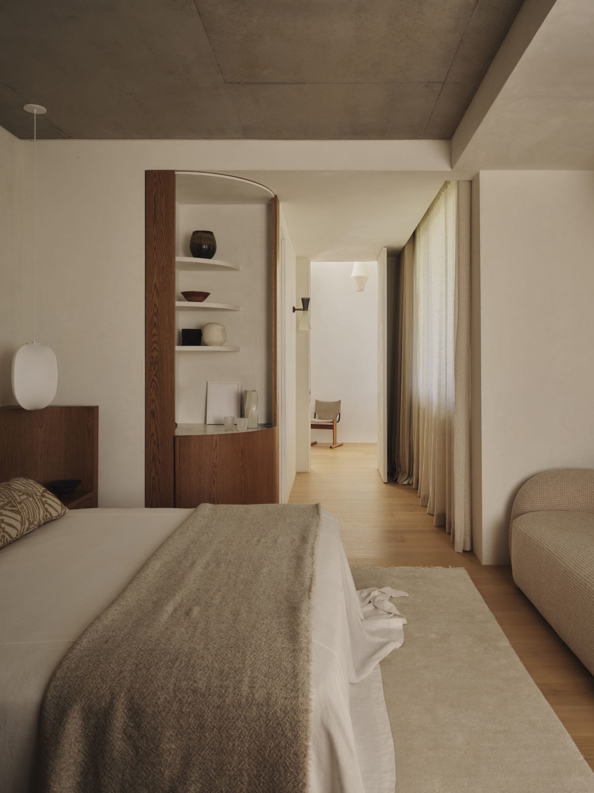 A calm and inviting bedroom with a bed, chair, and couch, decorated with wooden furniture and a white color scheme, with natural light streaming in through the window