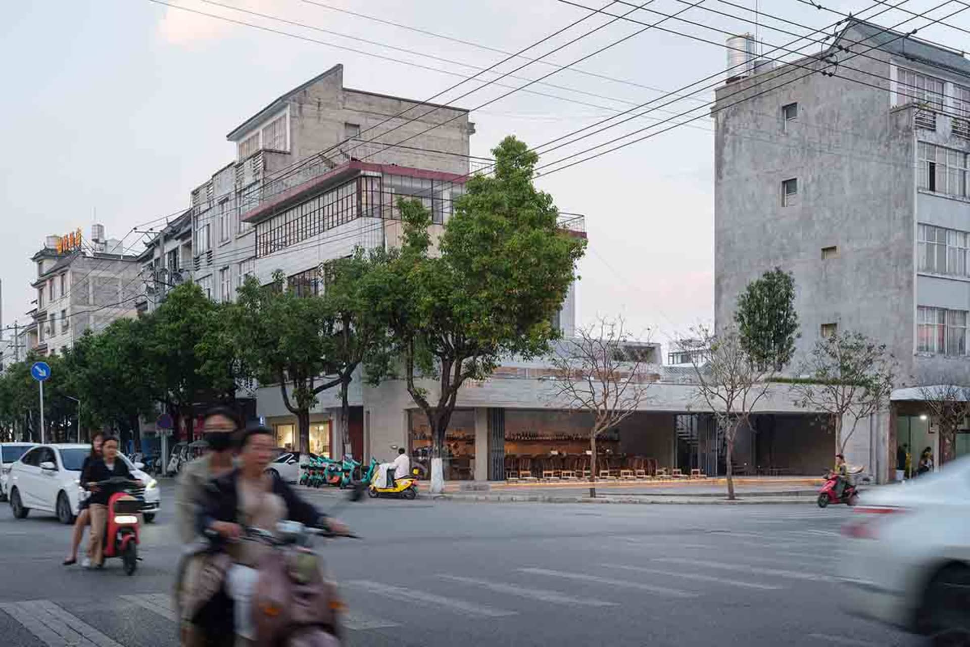 A busy city street with a tall building in the background, lined with trees and featuring motorcycles and pedestrians