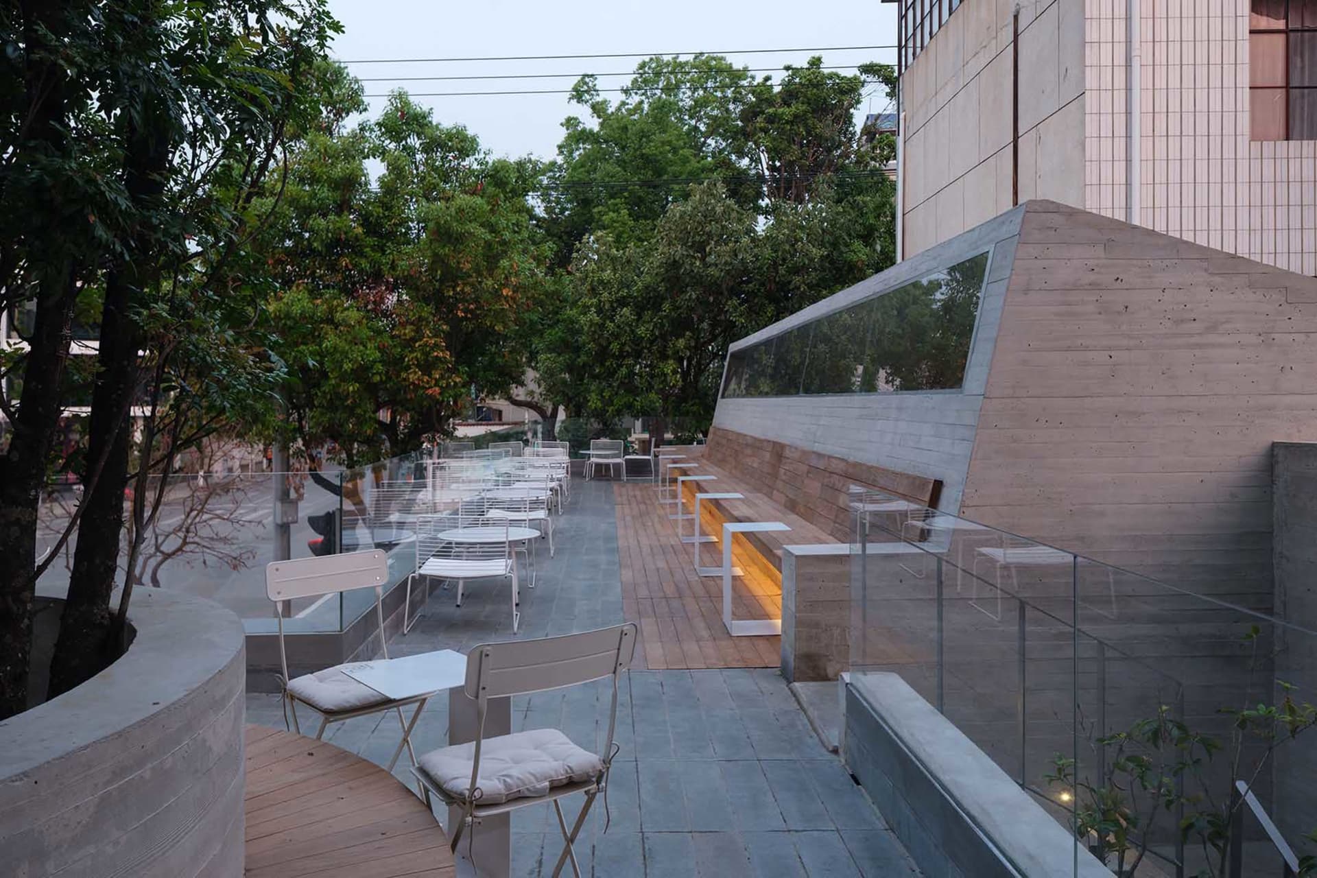 A modern outdoor seating area with a wooden deck, glass wall, and comfortable furniture, bathed in natural sunlight
