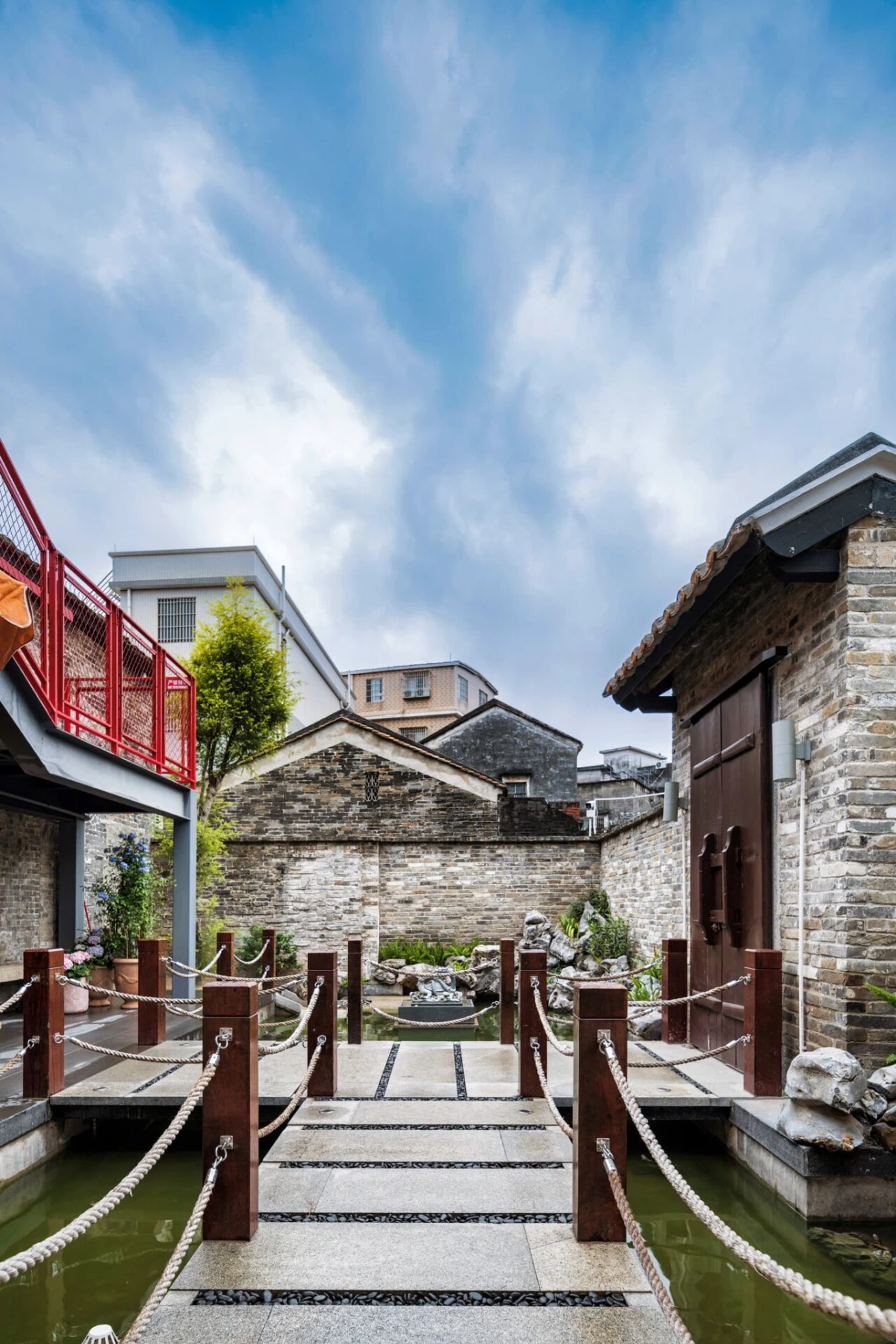 A serene courtyard with a stone building, brick wall, and wooden bridge, surrounded by potted plants and a pond, bathed in natural sunlight