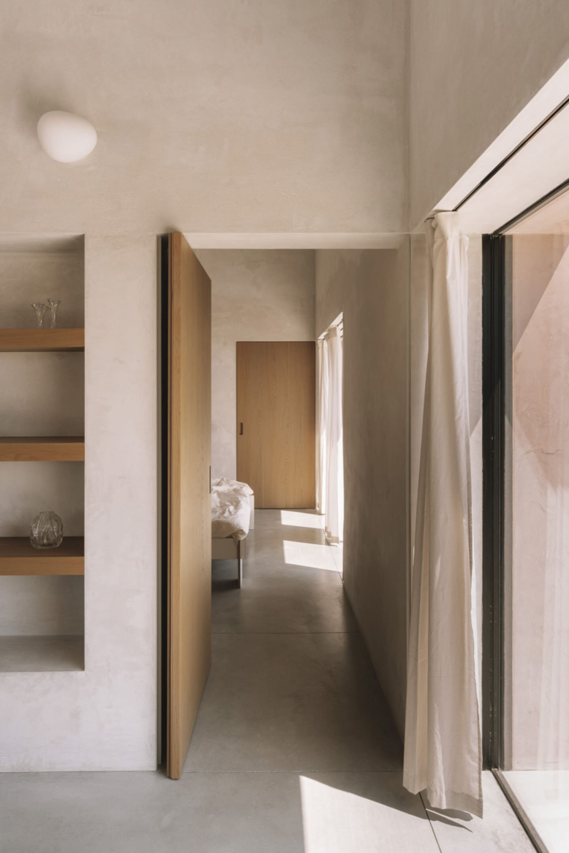 A cozy bedroom with a wooden floor, a white wall, a bed, a bookshelf, and a vase on the bookshelf, with natural light entering through an open door